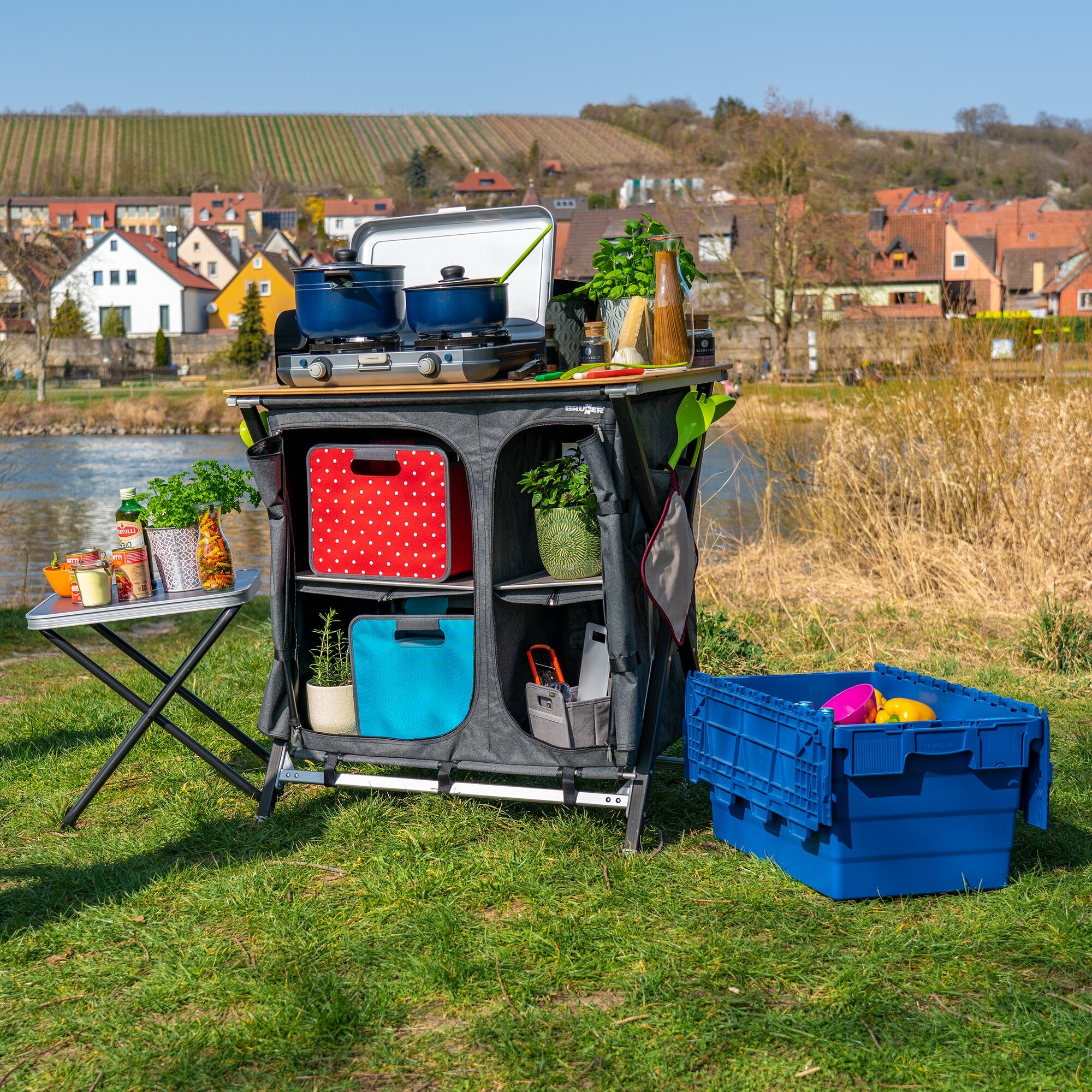Outdoor-Kochstation mit zwei blauen Töpfen auf Gasherd, auf tragbarem Tisch; grauer Mercury Cross Square Vielzweckschrank mit roter gepunkteter Fronttasche