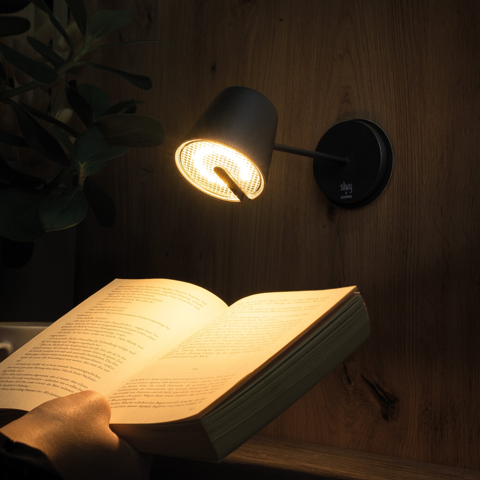 Schwarze Metall-Wandleuchte mit runder Wandhalterung und zylindrischem Leuchtenschirm beleuchtet warm, davor offenes Buch auf Holz