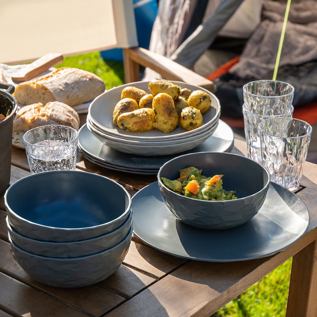 Graues CLUB GÖTEBORG Geschirrset aus Schüsseln und Tellern, mit Gläsern, auf einem Holztisch beim Camping, daneben Brot und Salat