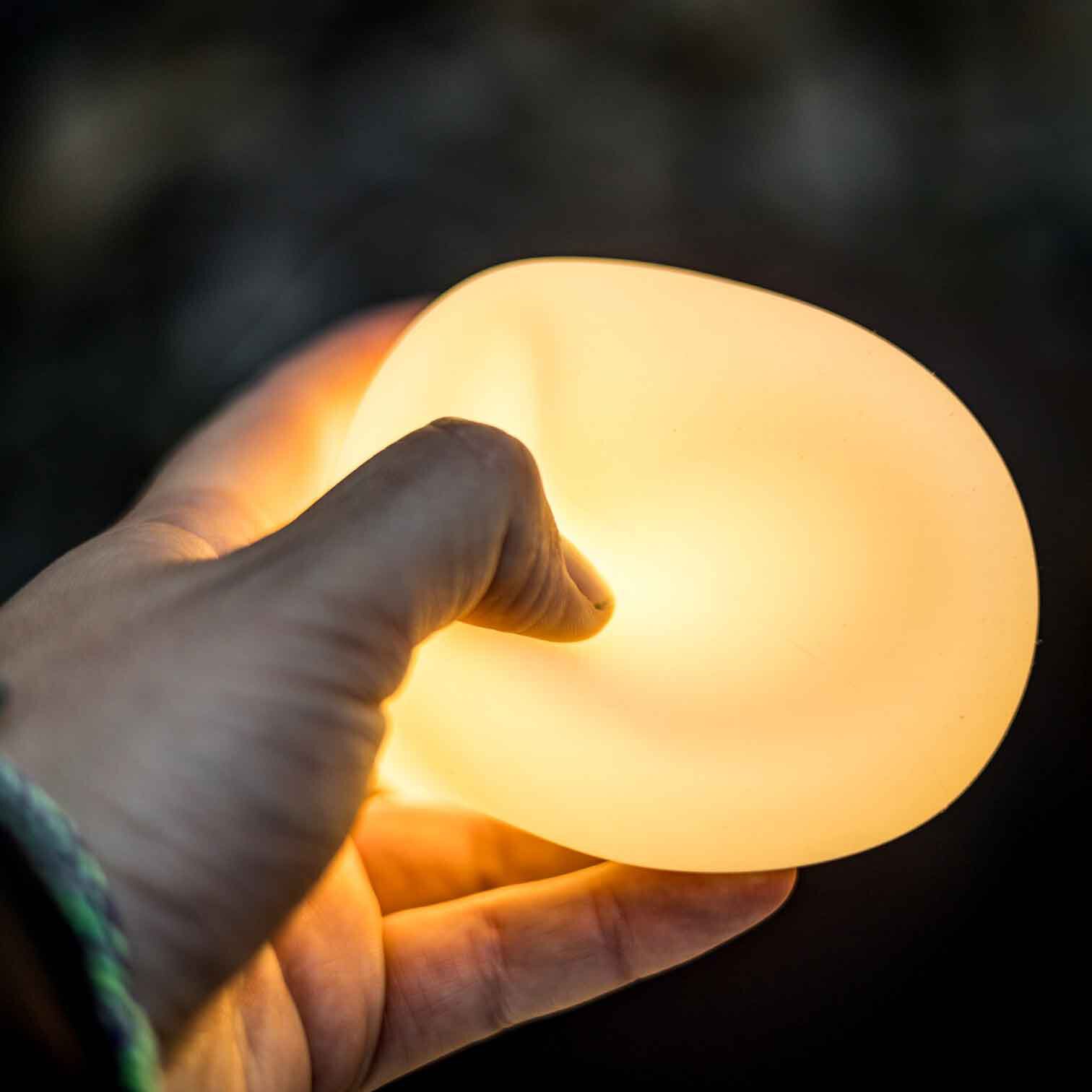 Nahaufnahme einer Hand, die eine runde Silikon-Campinglaterne hält, sie strahlt warmes Gelb aus vor dunklem Hintergrund