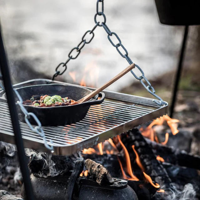 Schwenkbarer Edelstahlgrill über offenem Lagerfeuer, gusseiserne Pfanne mit Gemüse und Kochlöffel darauf, stabile.