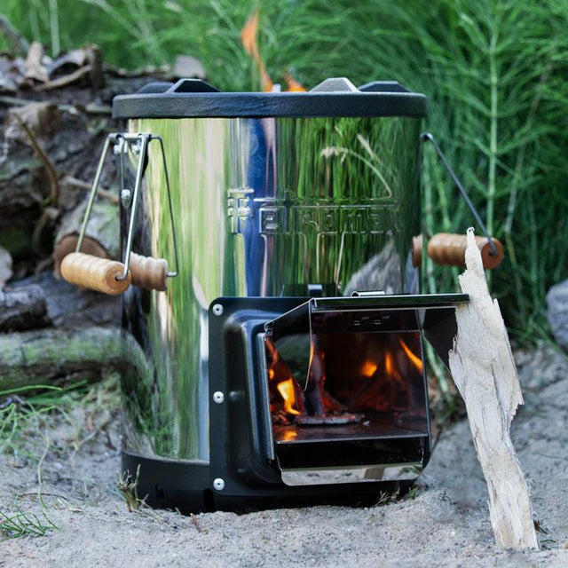 Runder Raketenofen aus glänzendem Edelstahl mit Holzgriff-Einsätzen, offener Feuerkammer mit sichtbarer Flamme, steht auf.