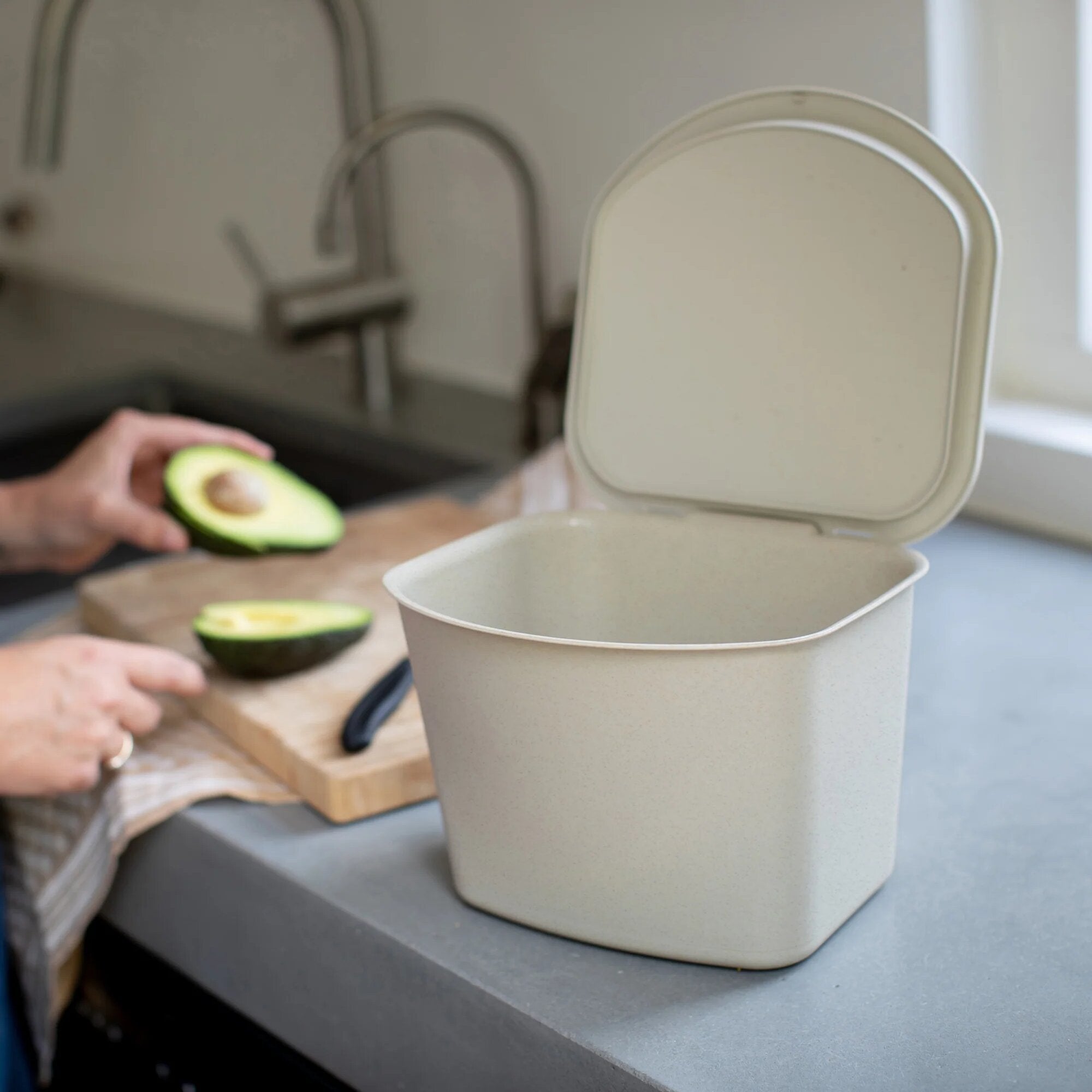 Sandfarbener Bioabfalleimer mit offenem Deckel in der Küche; im Vordergrund Avocadohälften auf einem Schneidebrett, Messer daneben