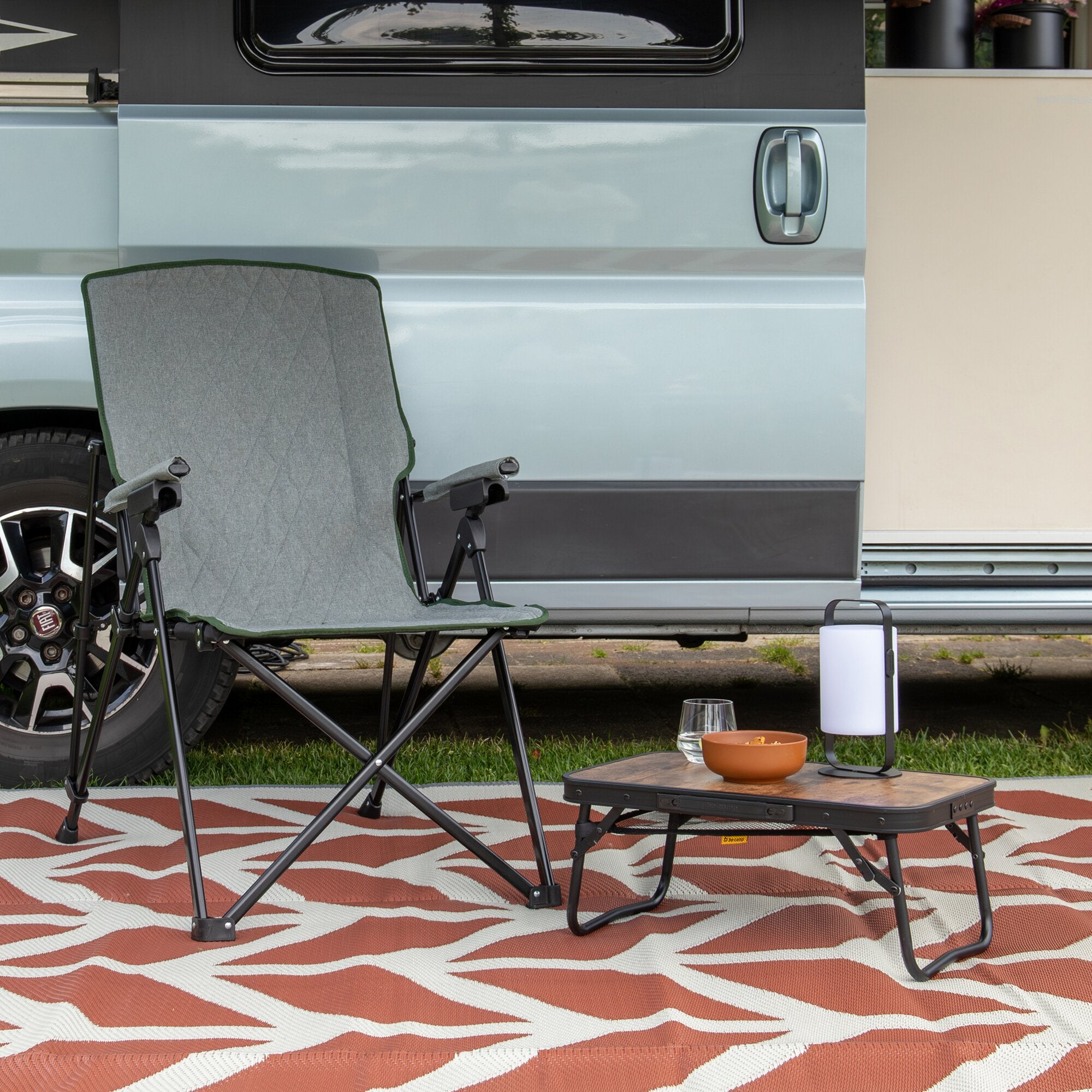 Grüner Stanwix Klappstuhl mit grauem Stoffsitz und schwarzem Metallrahmen, im Vordergrund auf rot-weißem Outdoor-Teppich vor einem Wohnmobil