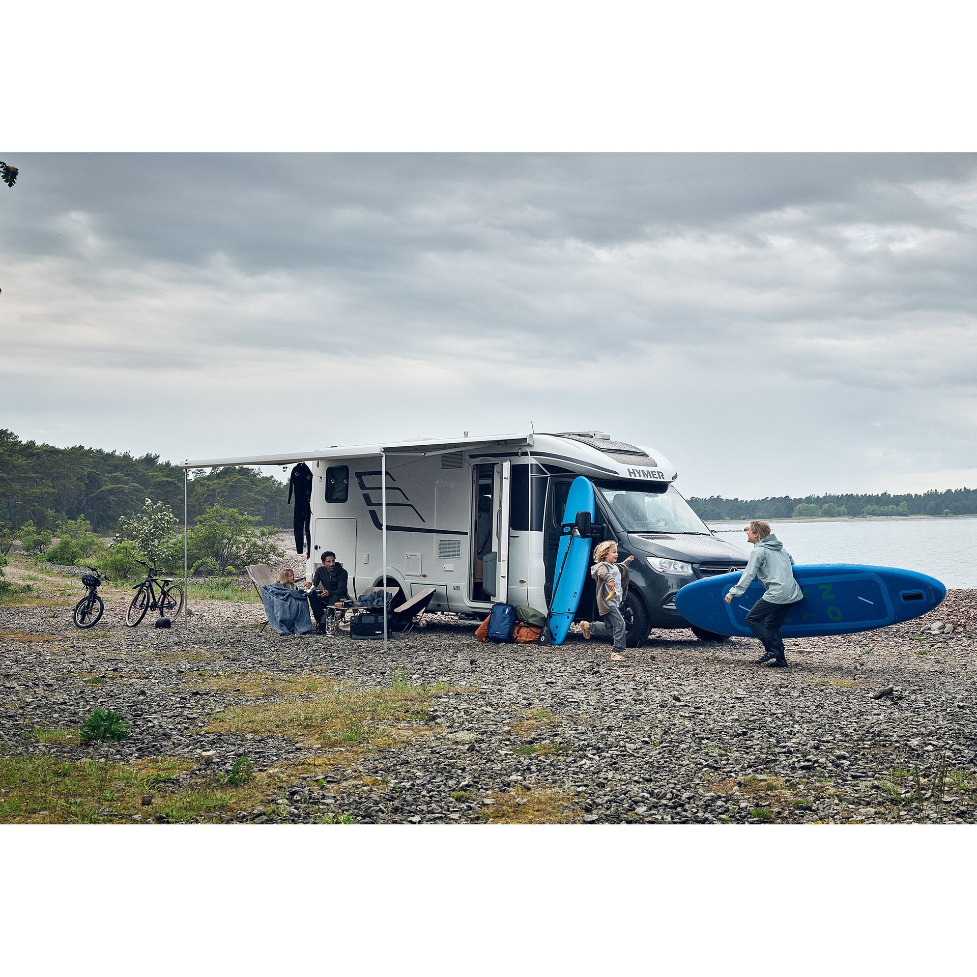Ausgefahrene Markise an einem weißen Wohnmobil am Kiesstrand, blaue Surfbretter stehen daneben, Fahrräder links im Bild, zwei Erwachsene und ein Kind im Camp, bewölkter Himmel