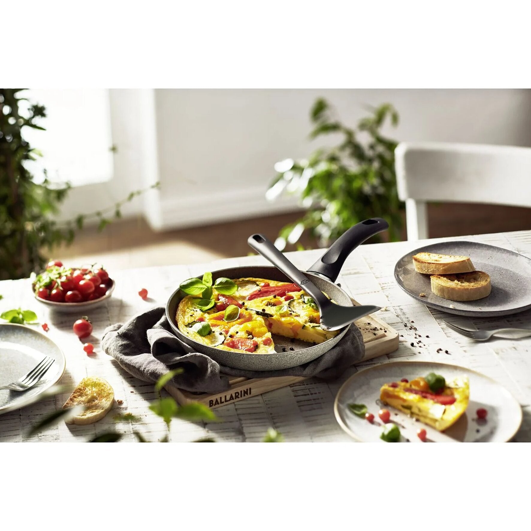 Schwarzer Wok-Wender aus dem Küchenhelfer Nero liegt auf einer Pfanne mit Quiche; daneben Brot, Tomaten und Basilikum, unscharfer Hintergrund