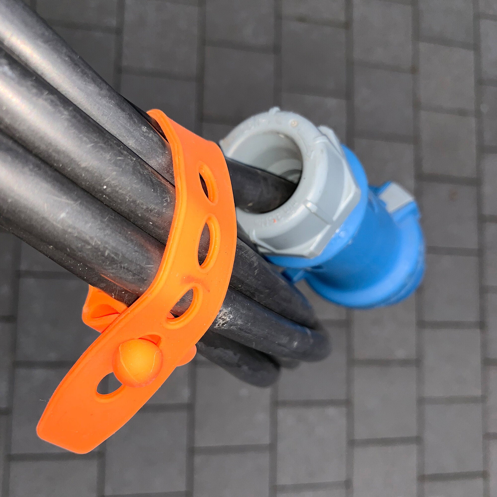 Detailaufnahme eines orangefarbenen Silikonbands mit Löchern, um einen schwarzen Rohrteil gelegt, grauer Kunststoffring und blauer Hintergrund sichtbar