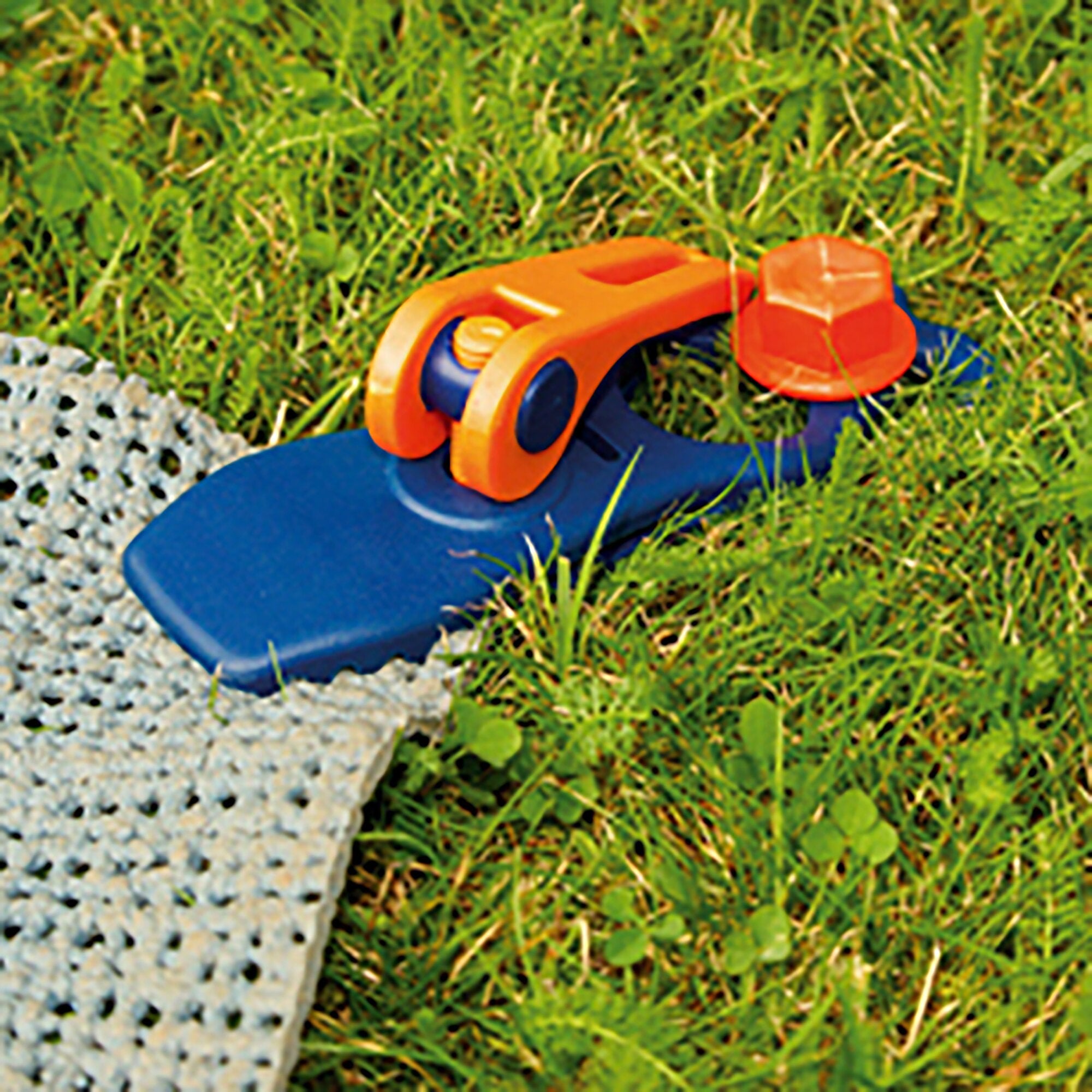 Nahaufnahme einer orangefarbenen Kunststoff-Schnellspannklemme mit blauem Innenteil und sichtbarer Schraube, auf grünem Gras neben blauem Untergrund