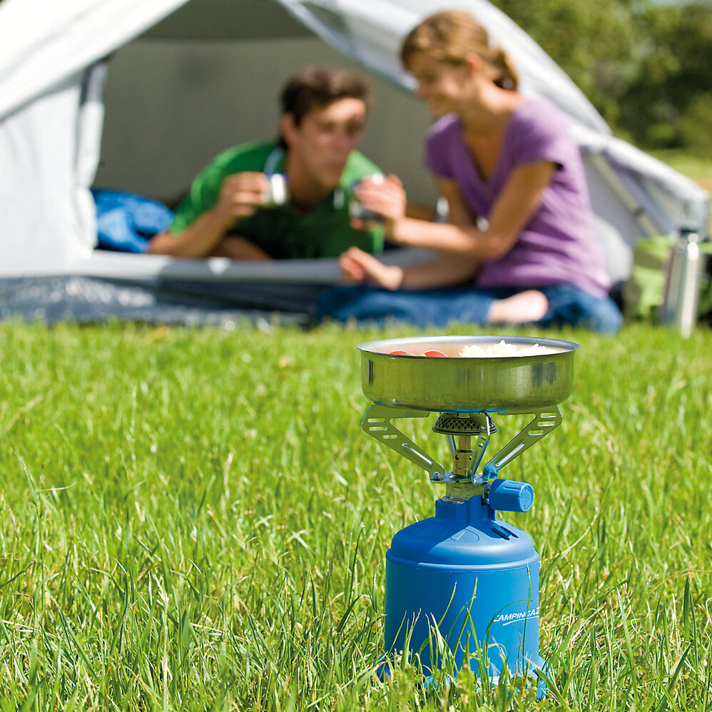 Campingkocher 206 S steht auf Gras, blauer Gaskartuschen-Kessel, Topf mit Essen darauf, im Hintergrund Zelt und zwei entspannte Personen