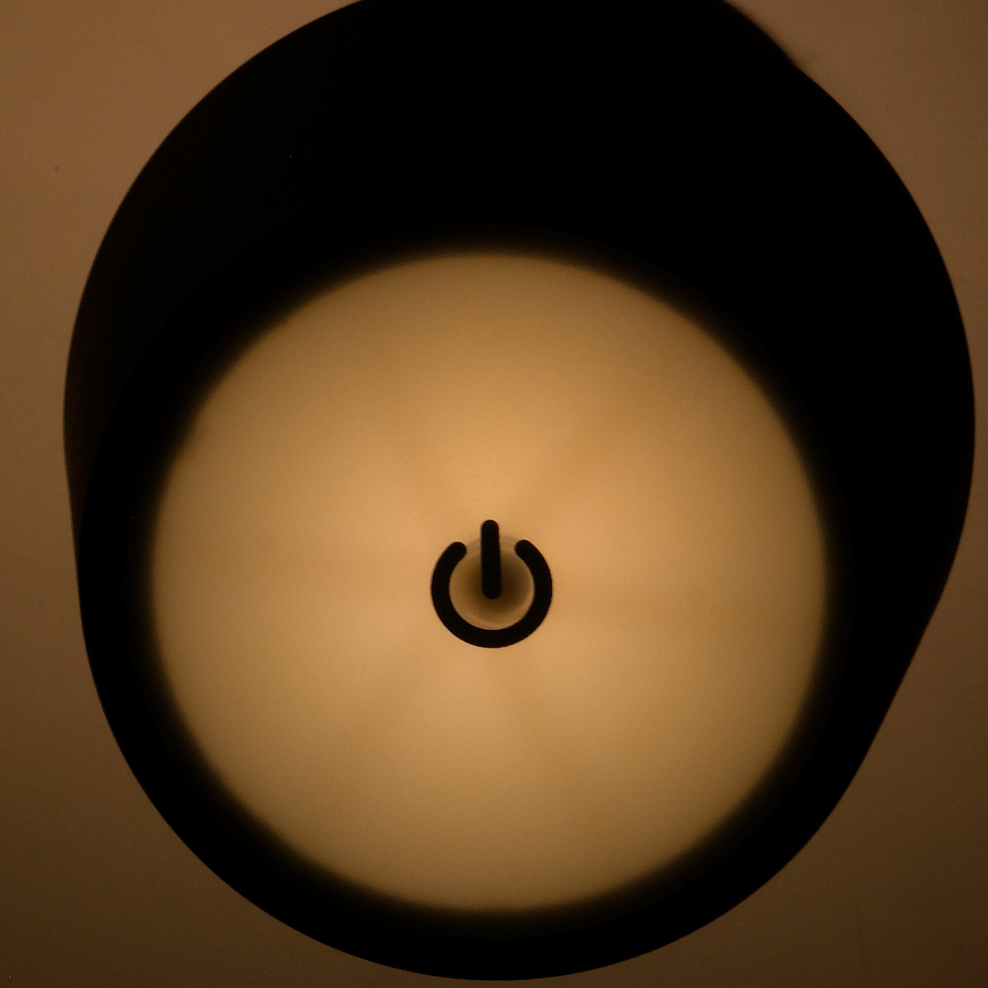 Nahaufnahme eines runden Ein-Aus-Knopf-Symbols (Power-Symbol) auf heller Fläche, umgeben von der dunklen Lampenoberfläche mit warmem Licht
