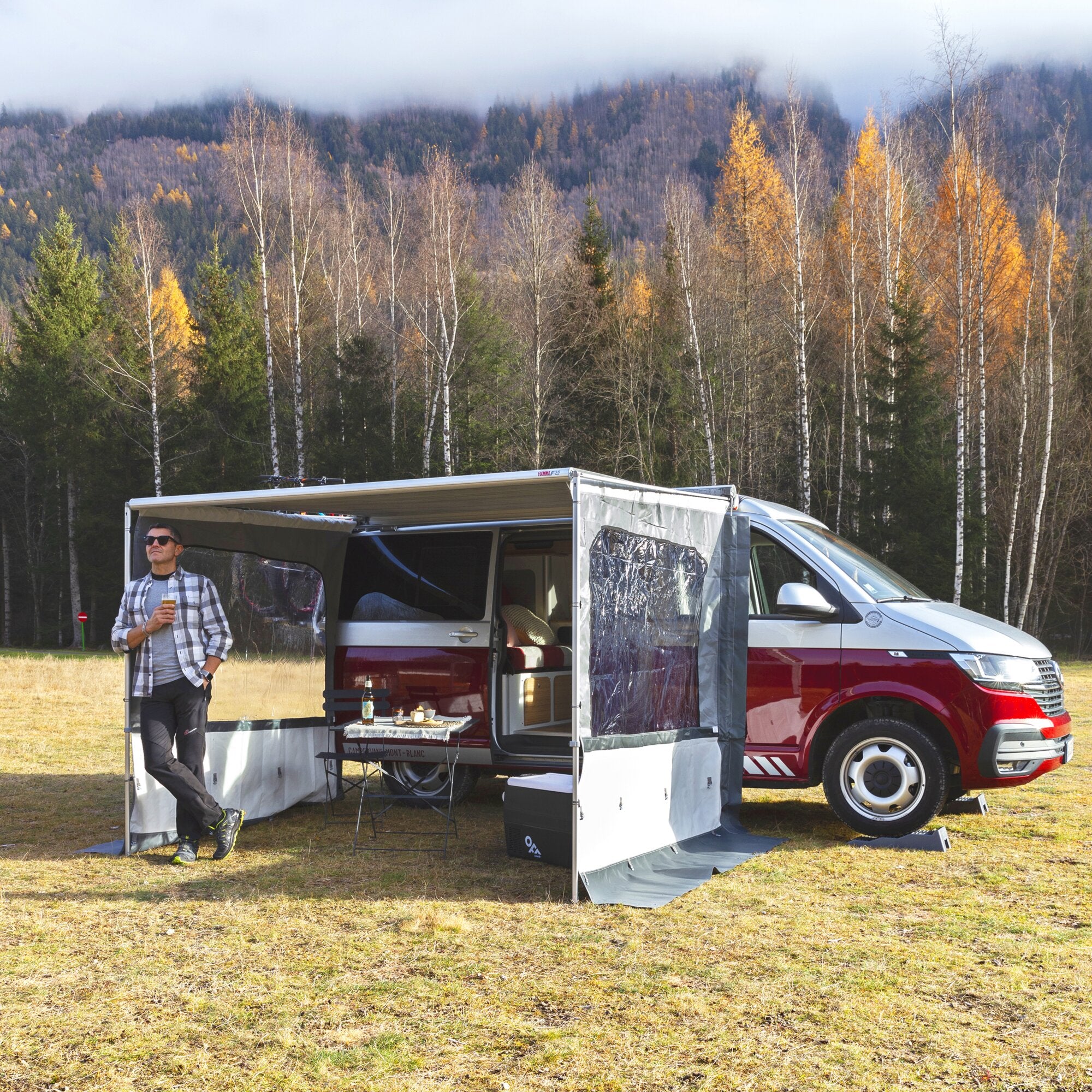 Offener Camper mit ausgeklappter Seitenwand Side W Pro F35 Van, Wohnraum mit Tisch und Stühlen, daneben eine Person mit Getränk, Wald im Hintergrund