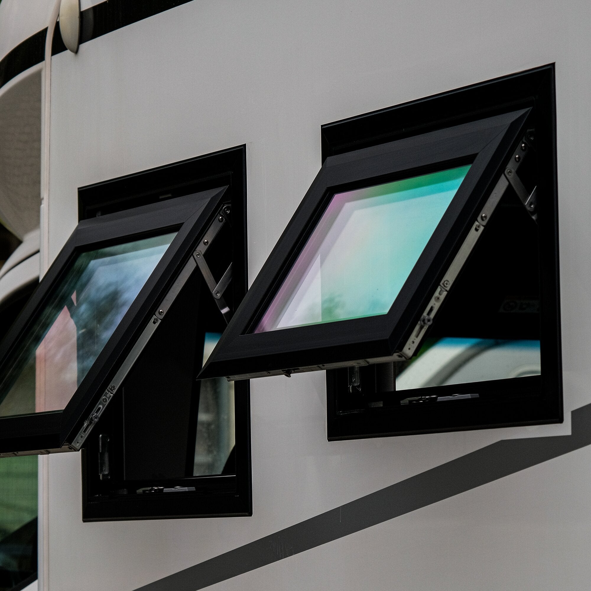 Nahaufnahme eines geöffneten Sicherheitsfensters mit schwarzem Metallrahmen, kippbaren Flügeln und reflektierendem Glas vor einer hellgrauen Wand