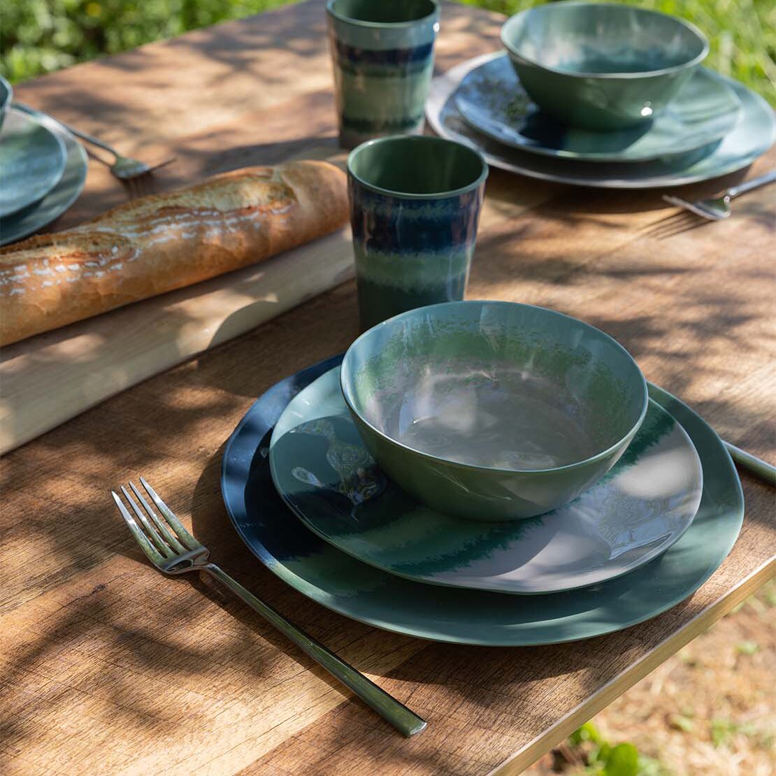Außentisch aus Holz mit buntem Halo 12-teiligem Geschirrset, grün-blau glasierte Schalen, Teller und Becher, daneben Brotlaib und Besteck im Schatten