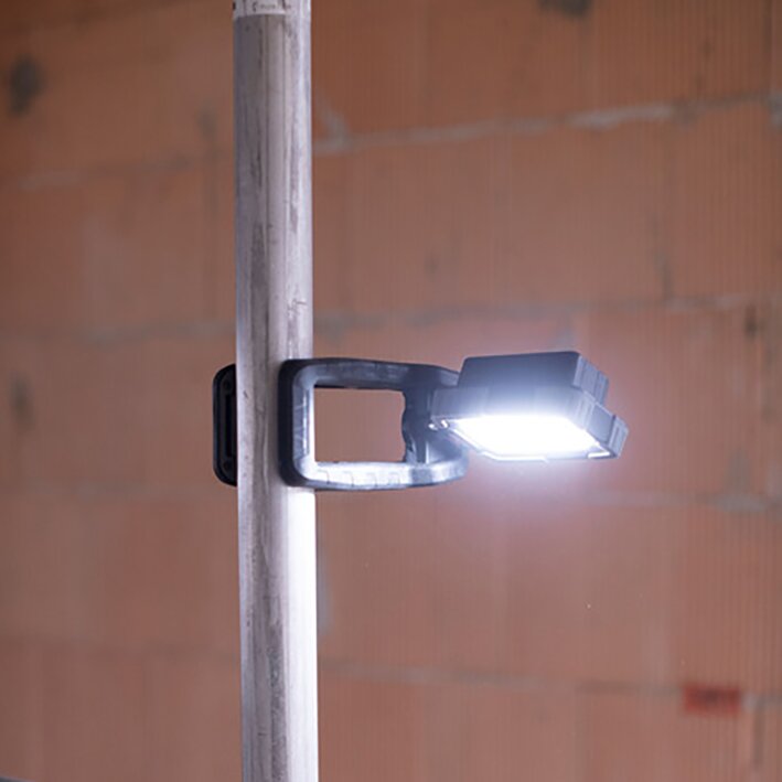 Klemmbarer mobiler LED-Arbeitsstrahler mit rechteckiger Leuchtfläche, an einer Stange befestigt, leuchtet hell, vor brauner Ziegelwand