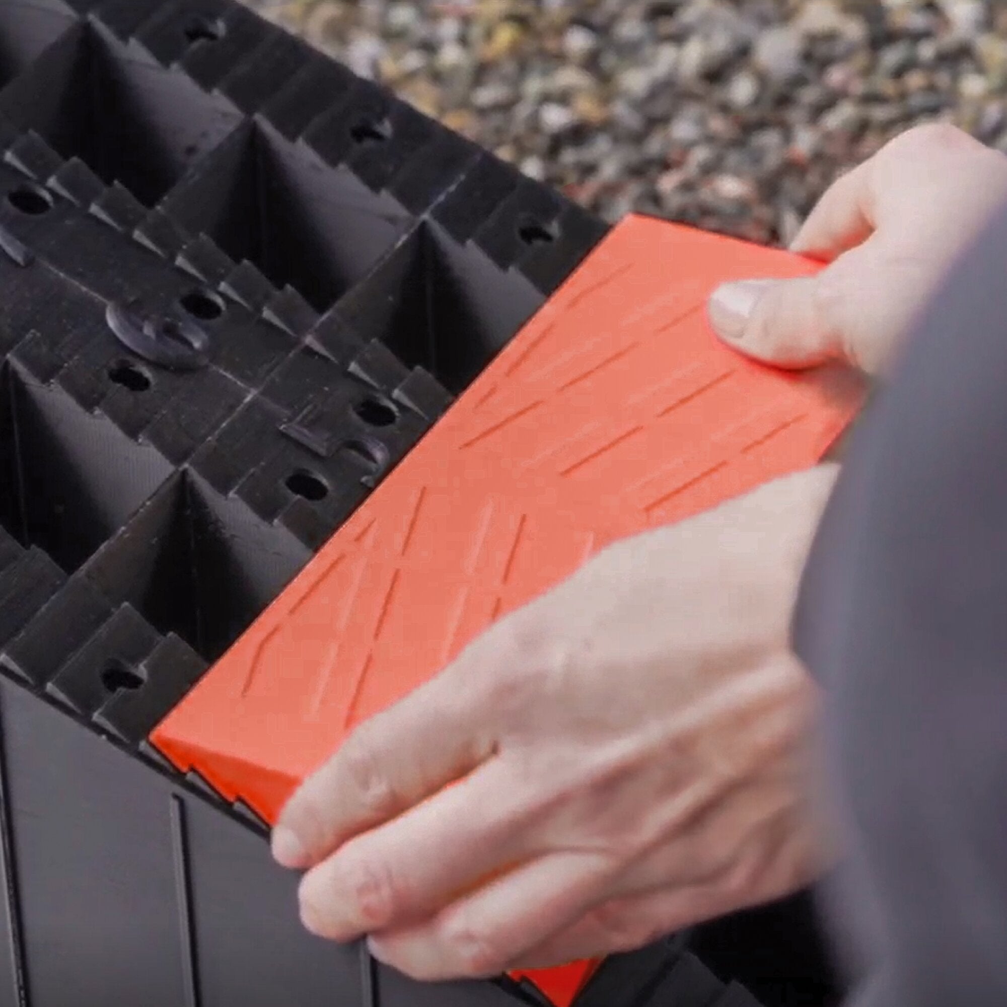 Nahaufnahme eines orangefarbenen Auffahrkeils, das von einer Hand gehalten wird; daneben schwarzes, mehrteiliges Blocksystem mit Löchern, Teil des LEVEL9 2er-Sets