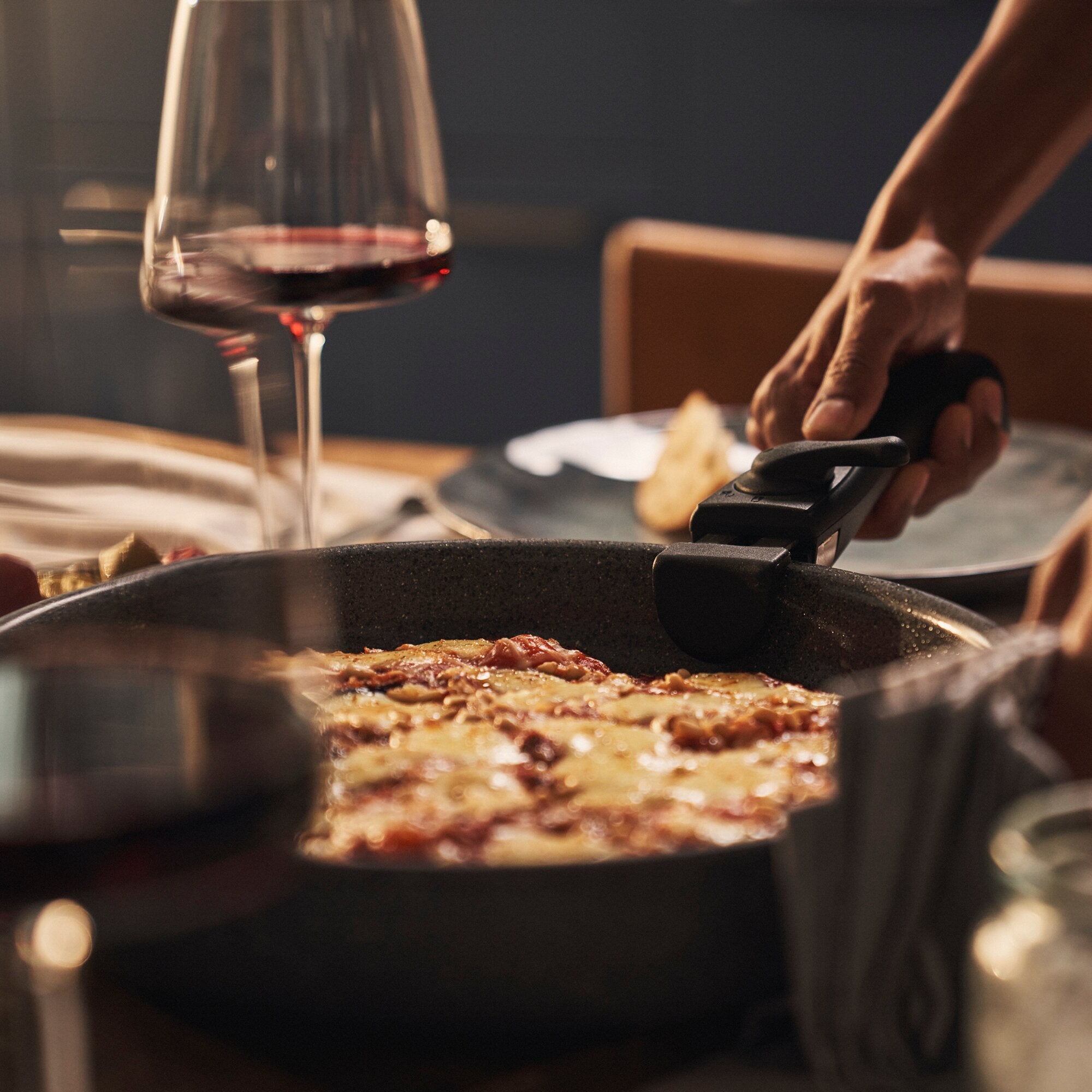 Große schwarze Bratpfanne mit angebratenem Fleisch auf dem Herd; daneben ein Glas Rotwein und eine Hand, die einen Küchengeräte-Griff hält