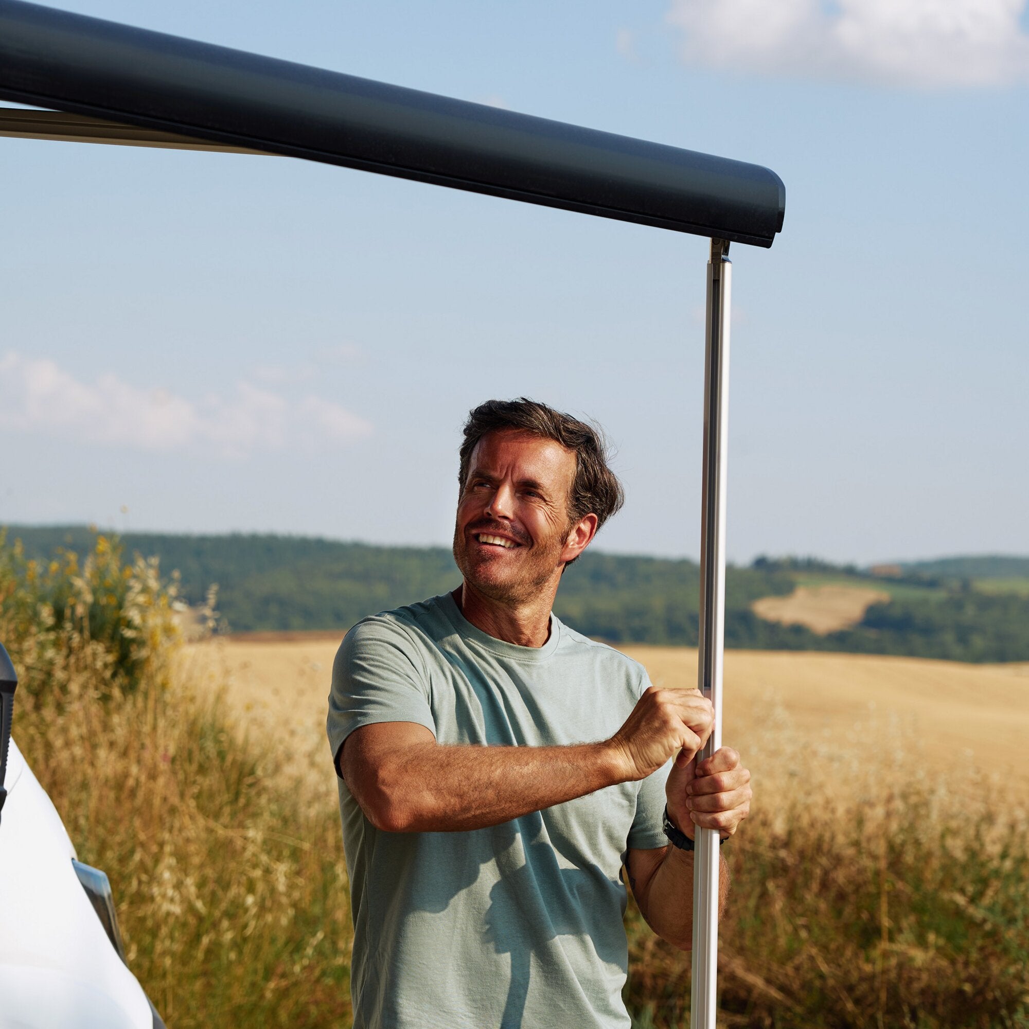 Mann hält im Freien eine schwarze Markise F80 mit rollendem Gehäuse und Stützrohr, Himmel und weite Landschaft im Hintergrund