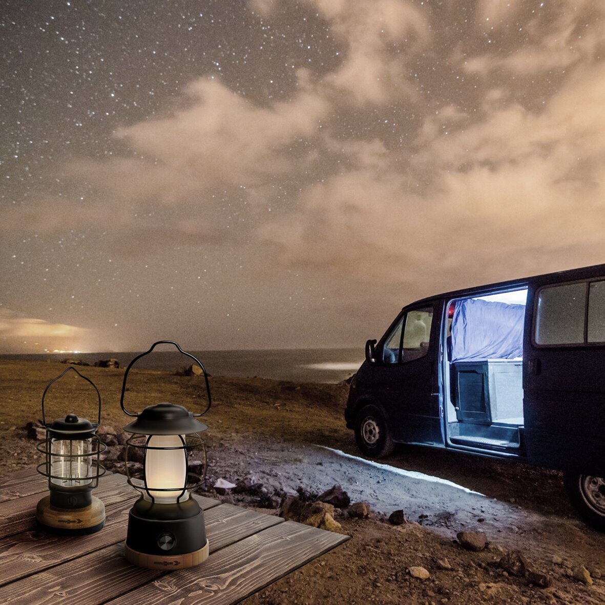 Zwei Akku-Campinglampen Laterne Lyss stehen auf einer Holzplanke neben einem offenen Camping-Van, nächtlicher Sternenhimmel am Meer