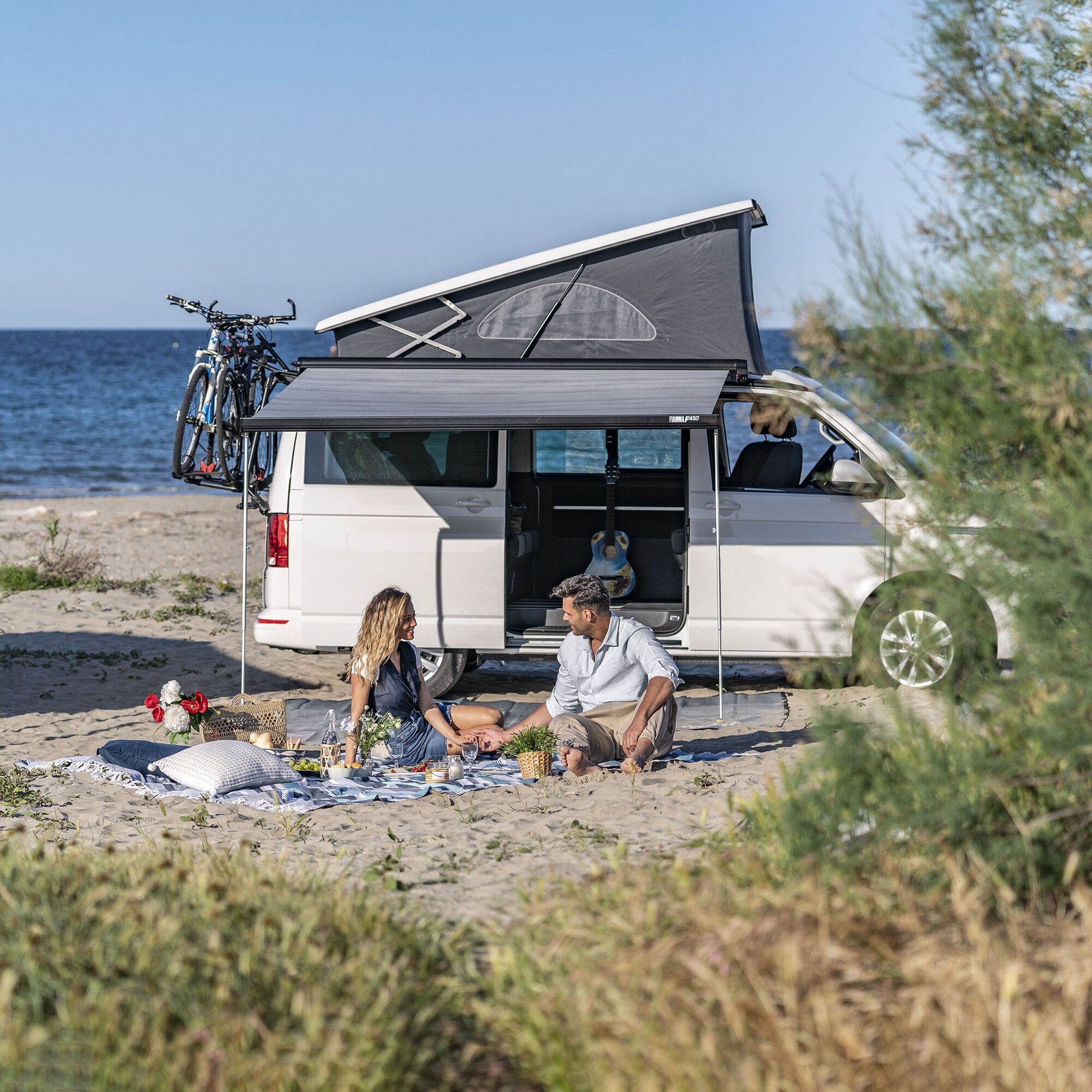 Weißer VW T5/T6 Multivan mit schwarzer F45s Markise seitlich am Wagen ausgeklappt; Fahrräder am Heckträger, Paar genießt Picknick am Strand