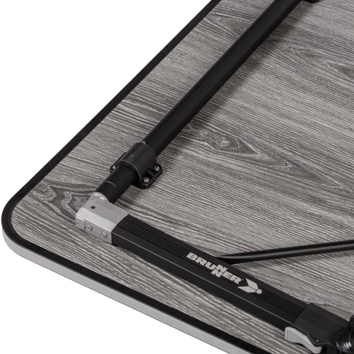 Detailaufnahme einer Campingtischplatte mit grauer Holzoptik und schwarzem Rand, sichtbarer schwarzer Metallstab mit Verbindungsbeschlag und Brunner-Logo am Rahmen