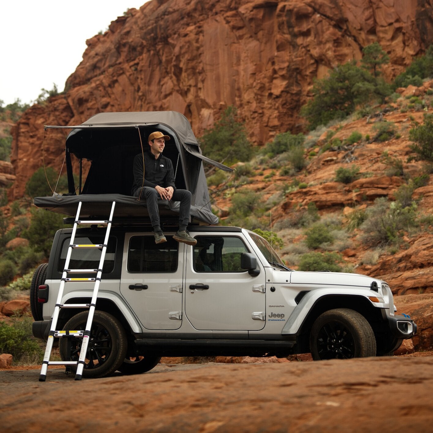 Auf einem Jeep Wrangler steht ein aufgeklapptes Dachzelt Roof Space 2 mit Leiter, ein Mann sitzt am Zeltrand vor rotbraunen Felsen