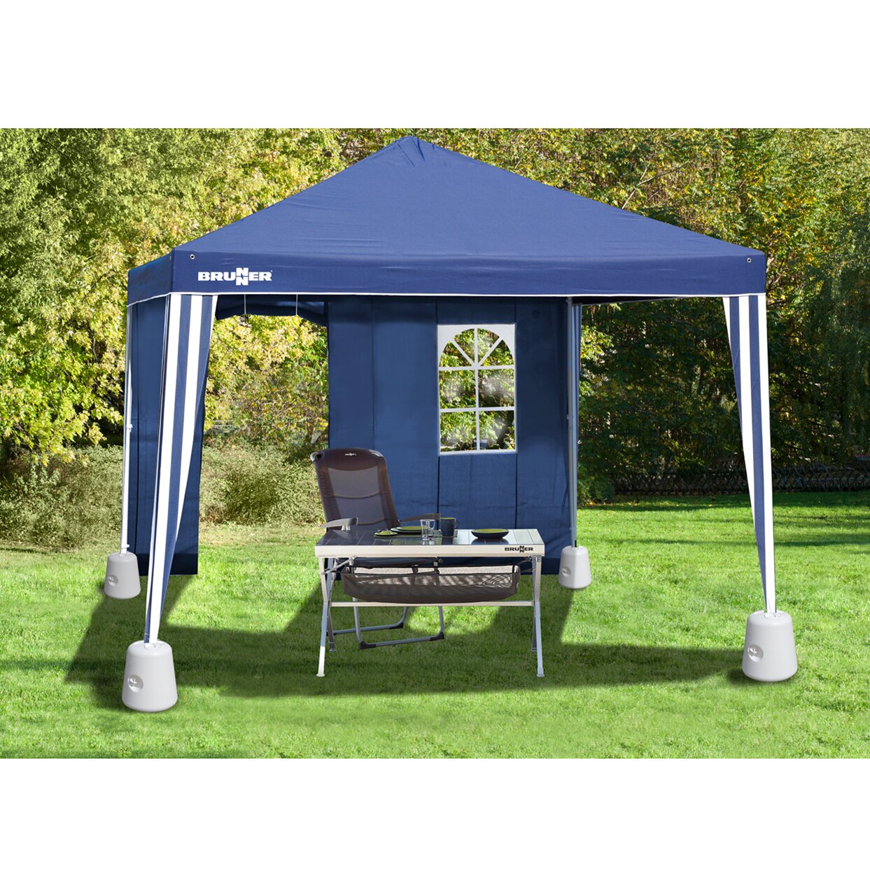 Blauer Pavillon mit weißem Gestell und runden Bodengewichten an den Beinen steht auf grünem Rasen; davor Tisch und Stuhl, Gartenhintergrund