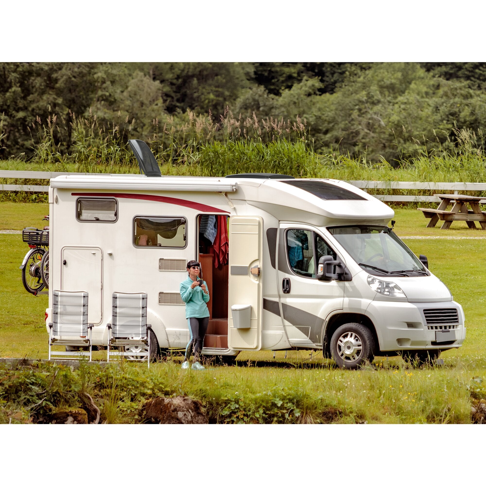Weißes Wohnmobil mit offener Tür; auf dem Dach sichtbar eine Dachhaube/Lüfter, dahinter Gras, Bäume und eine Person in Türkis neben dem Fahrzeug