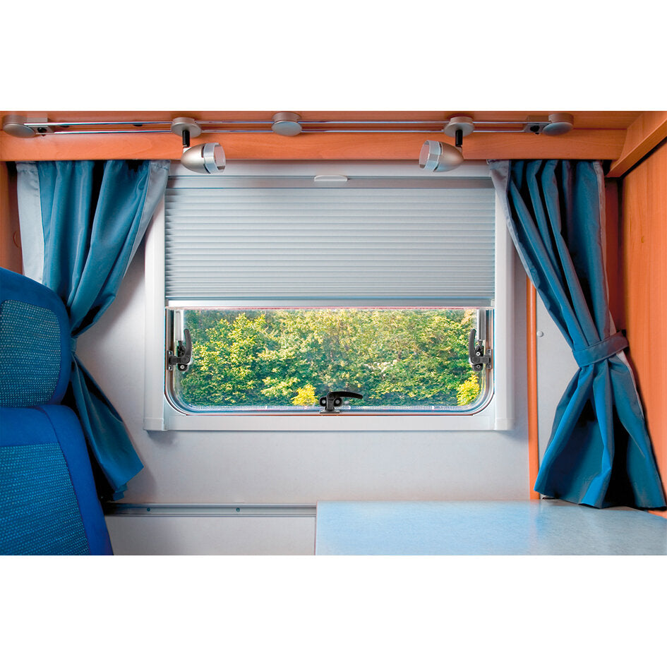 Innenraum eines Wohnmobils mit hellgrauen REMIflair IV Rollo über dem Fenster, blaue Vorhänge seitlich, orange Holzdekor, Blick nach draußen aufs Grün