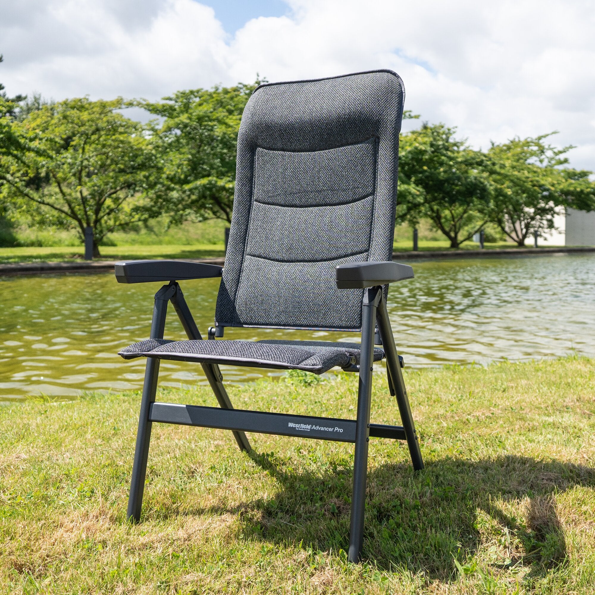 Rückenlehne eines dunkelgrauen, gewebten Campingstuhls Advancer Pro mit Armlehnen vor Gras, See und Bäumen in einer Outdoor-Szene