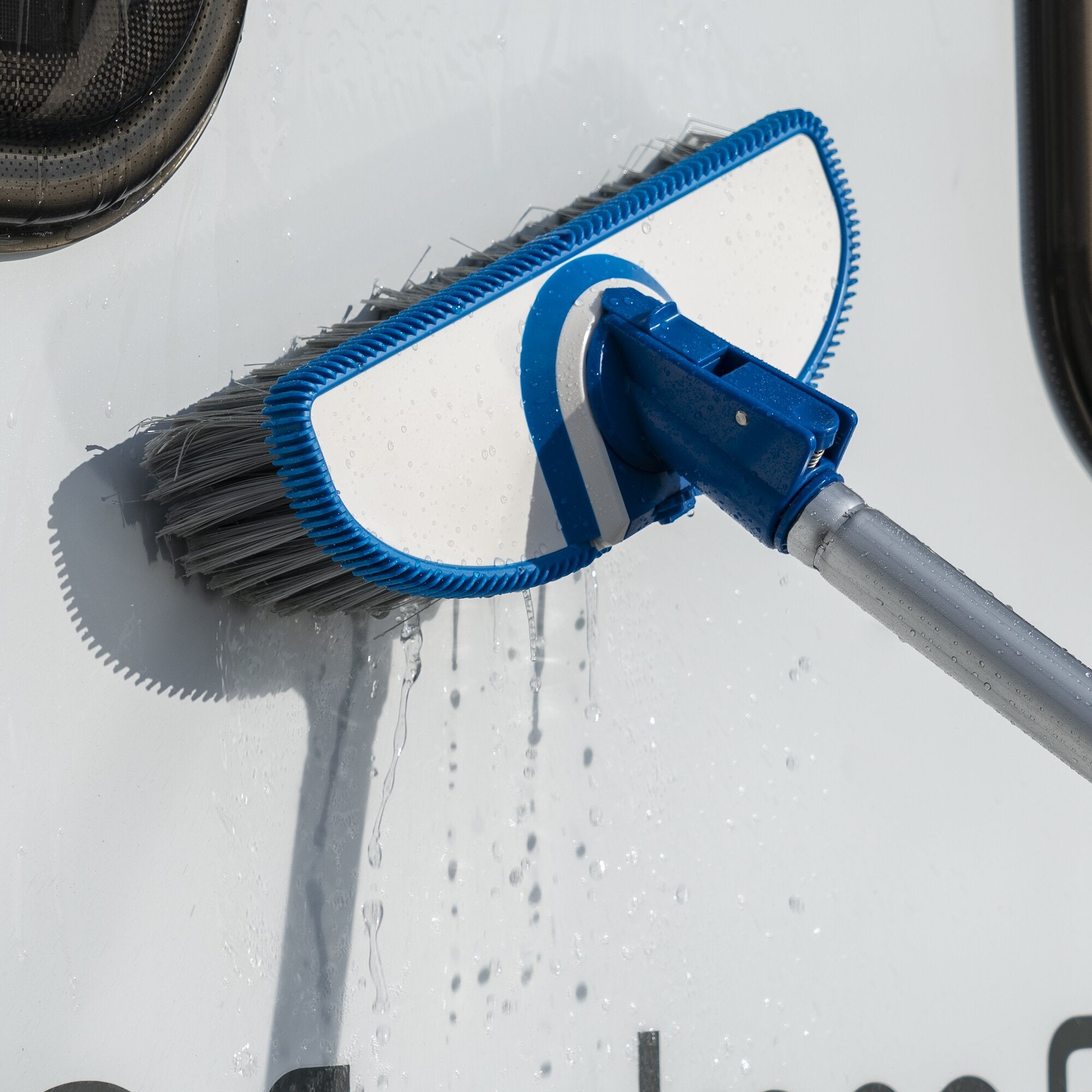 Nahaufnahme eines blauen, ovalen Reinigungswerkzeugs mit einer Borstenkante und grauem Griff, Wassertröpfchen auf einer glatten weißen Fläche
