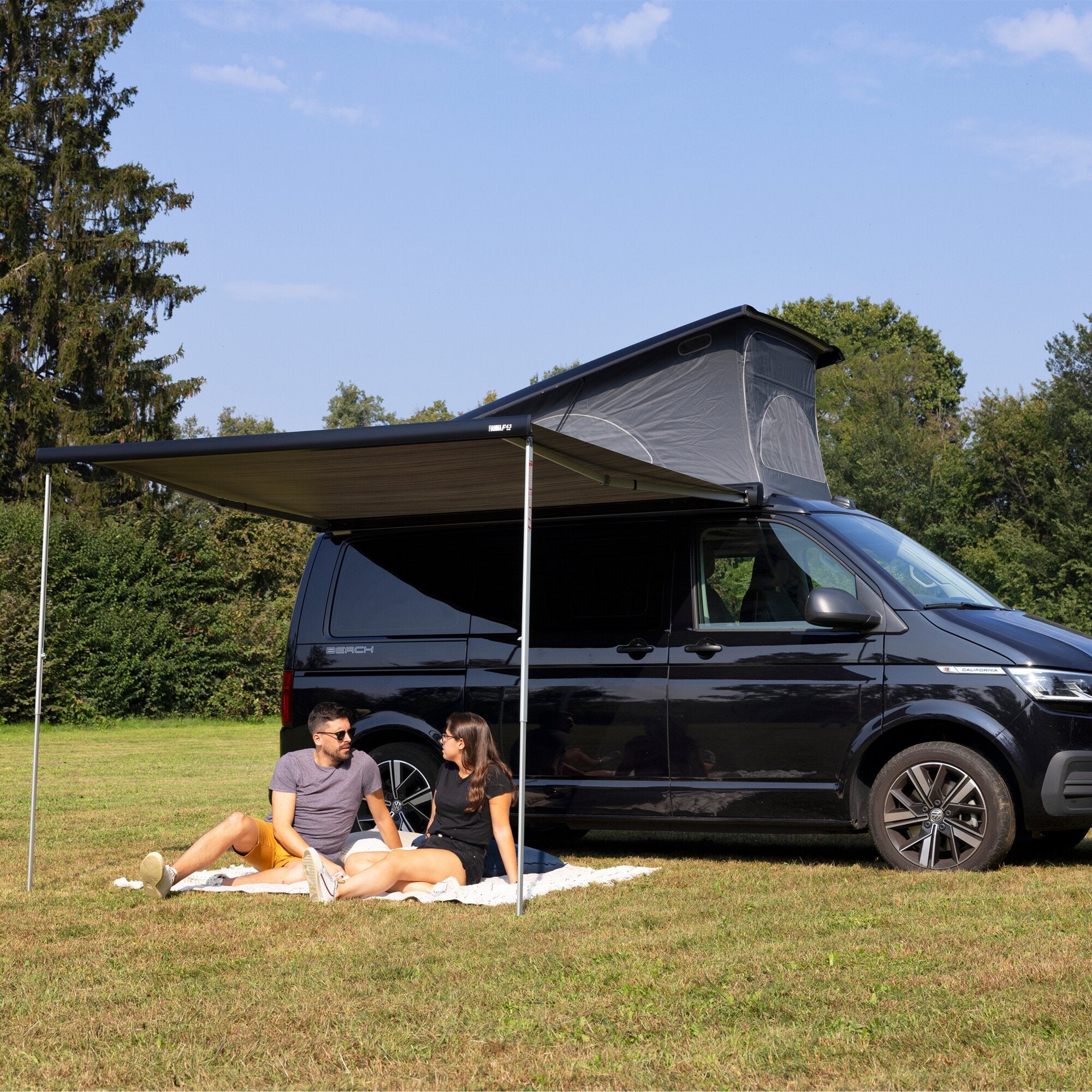 Außenansicht eines schwarzen Campervans mit einer ausgefahrenen Markise F43van 270 cm und Dachzelt, davor sitzen zwei Personen auf Decke im Gras