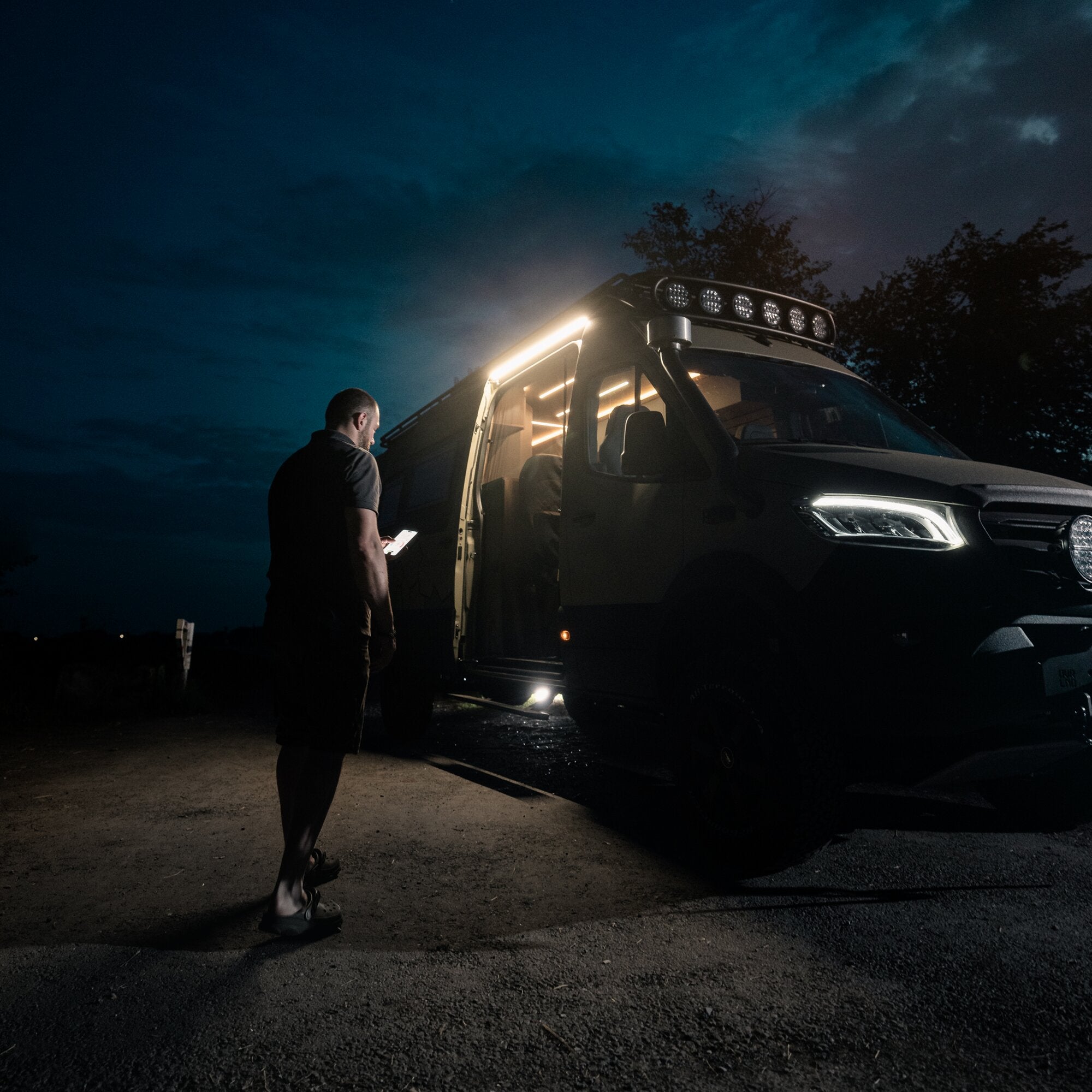 Nachtaufnahme eines schwarzen Transporters mit offener Fahrertür; Innenraum beleuchtet, Dachleiste mit LED-Modulen; Mann schaut auf Tablet