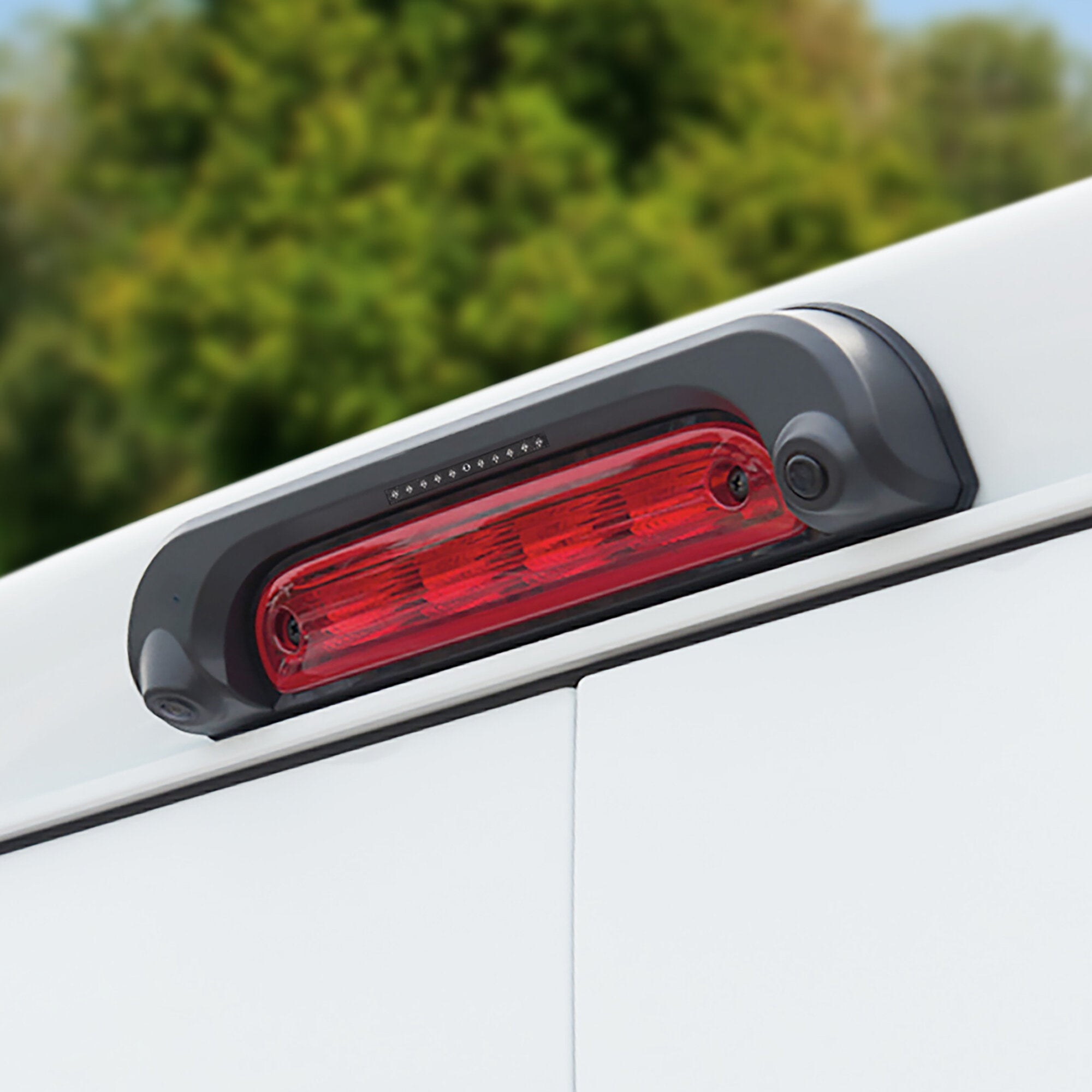 Nahaufnahme eines weißen Fahrzeughecks mit integrierter Rückfahrkamera im grauen Gehäuse neben dem roten Rücklicht, unscharfer grüner Hintergrund