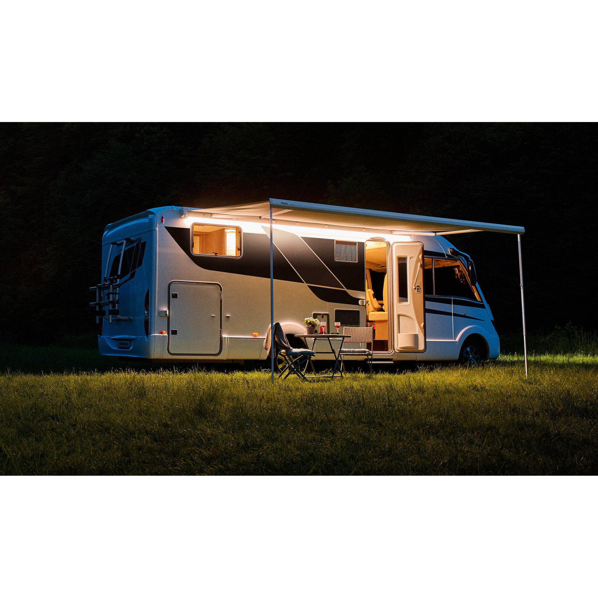 Nachtaufnahme eines Wohnmobils mit Seitenteil; unter dem Vorzelt leuchtet ein LED-Lichtstreifen und beleuchtet Tisch mit zwei Stühlen, Wald im Hintergrund