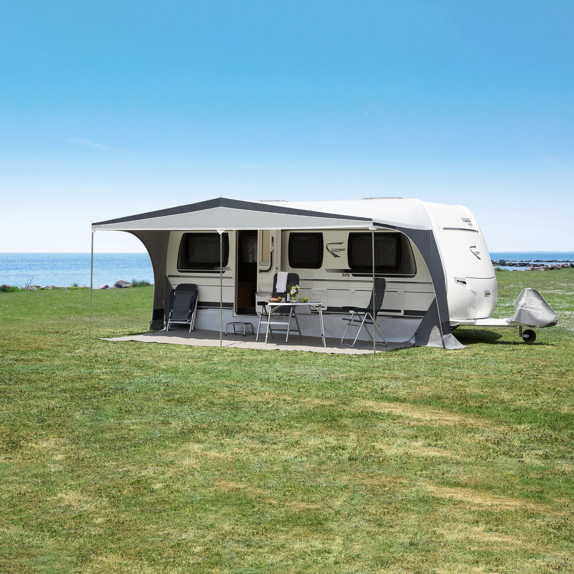 Wohnwagen mit ausgefahrener Markise, Outdoor-Tisch und Stühle, grüne Wiese, Meer im Hintergrund, blauer Himmel