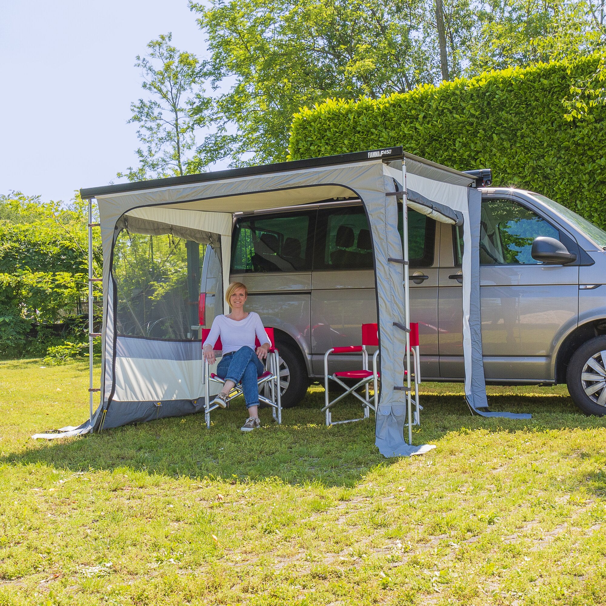 Wohnmobil mit seitlich ausgefahrener Markise aus grauem Stoff, davor zwei rote Klappstühle, Grasboden, grüne Hecke und Bäume im Hintergrund