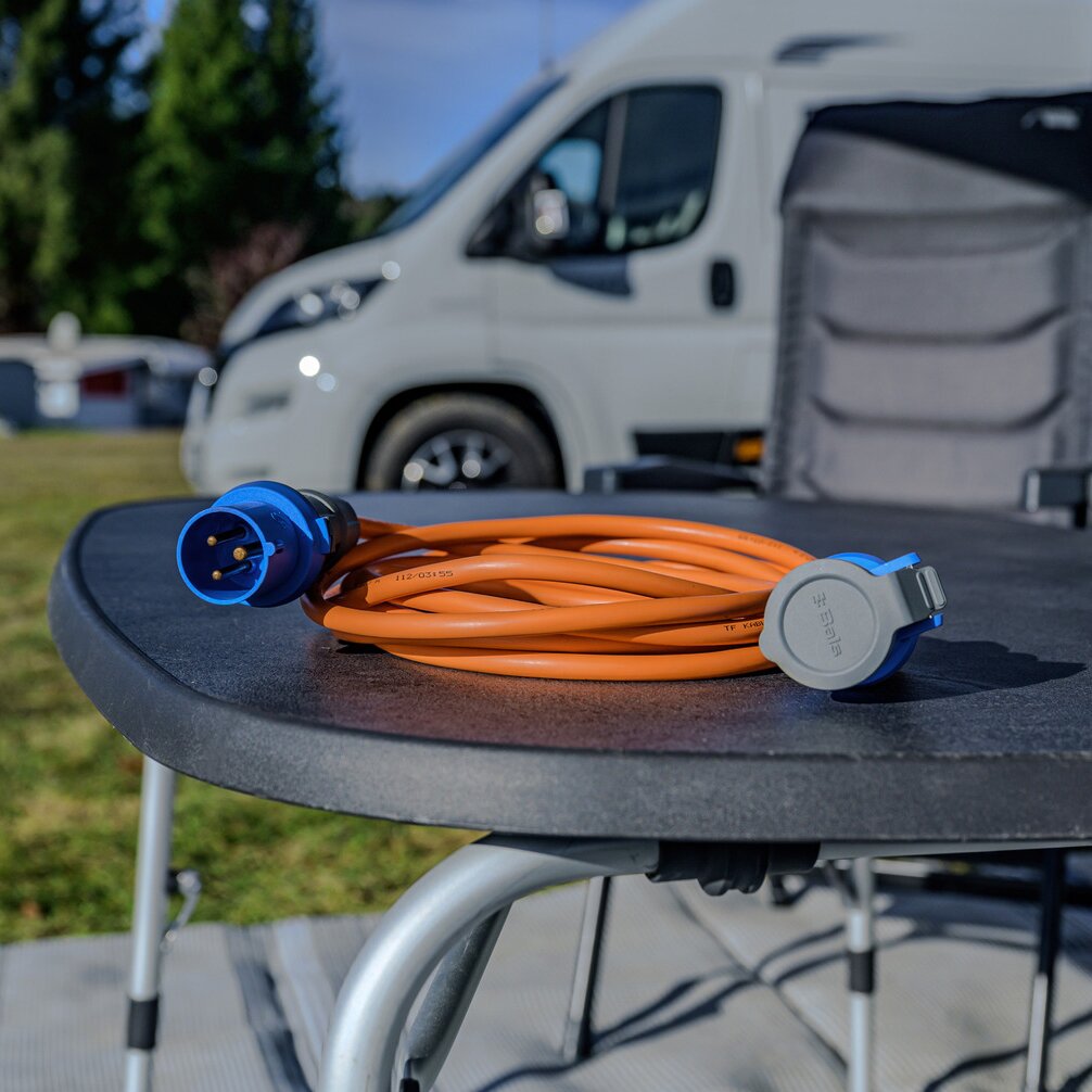 Orange CEE-Verlängerungskabel aufgewickelt, blauer Rundstecker sichtbar, liegt auf einem runden Tisch; Wohnmobil im Hintergrund