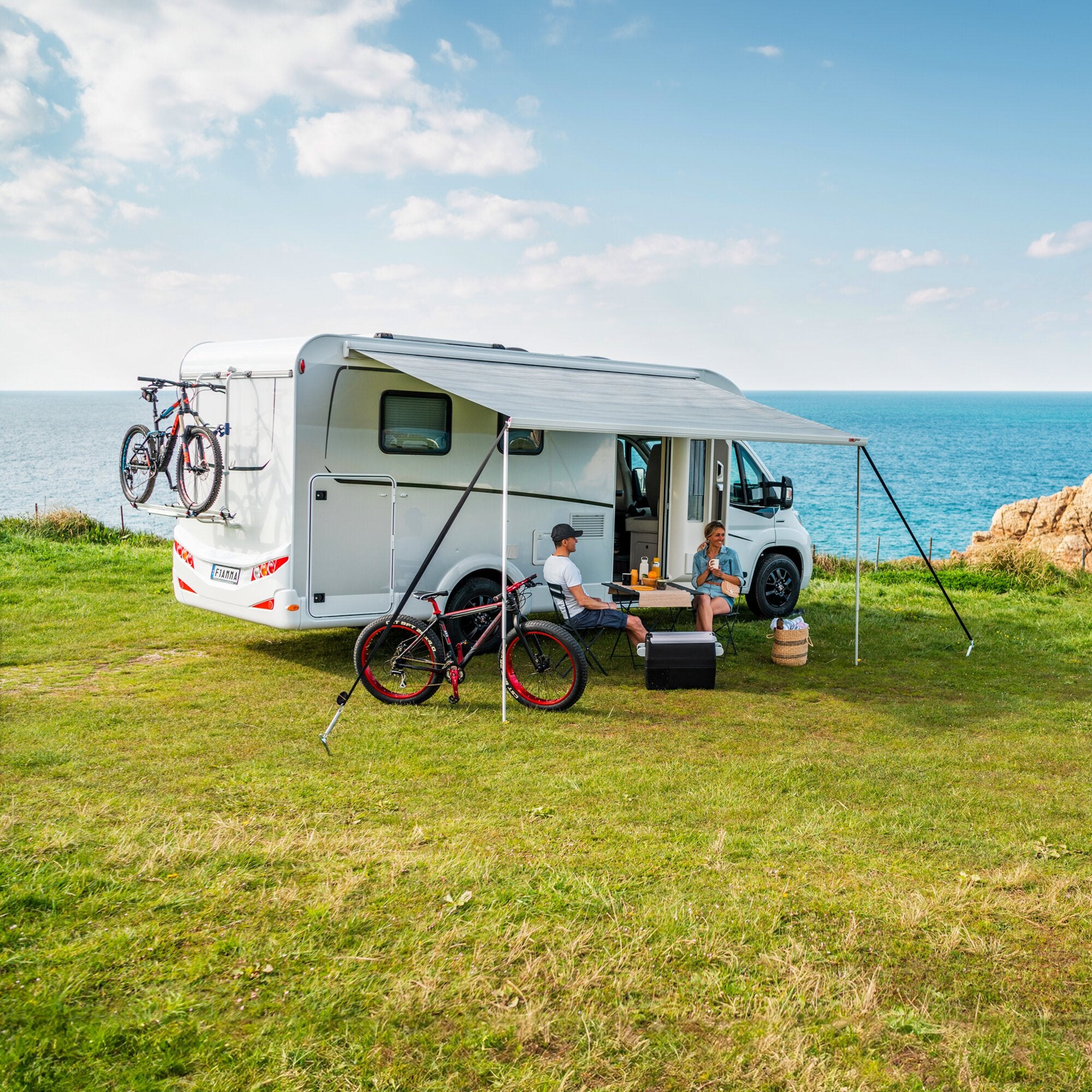 Weißes Wohnmobil am Meer mit ausgezogener Markise F45 in polarweiß, Fahrräder am Heckträger, zwei Personen sitzen an einem Tisch mit Getränken auf grünem Gras