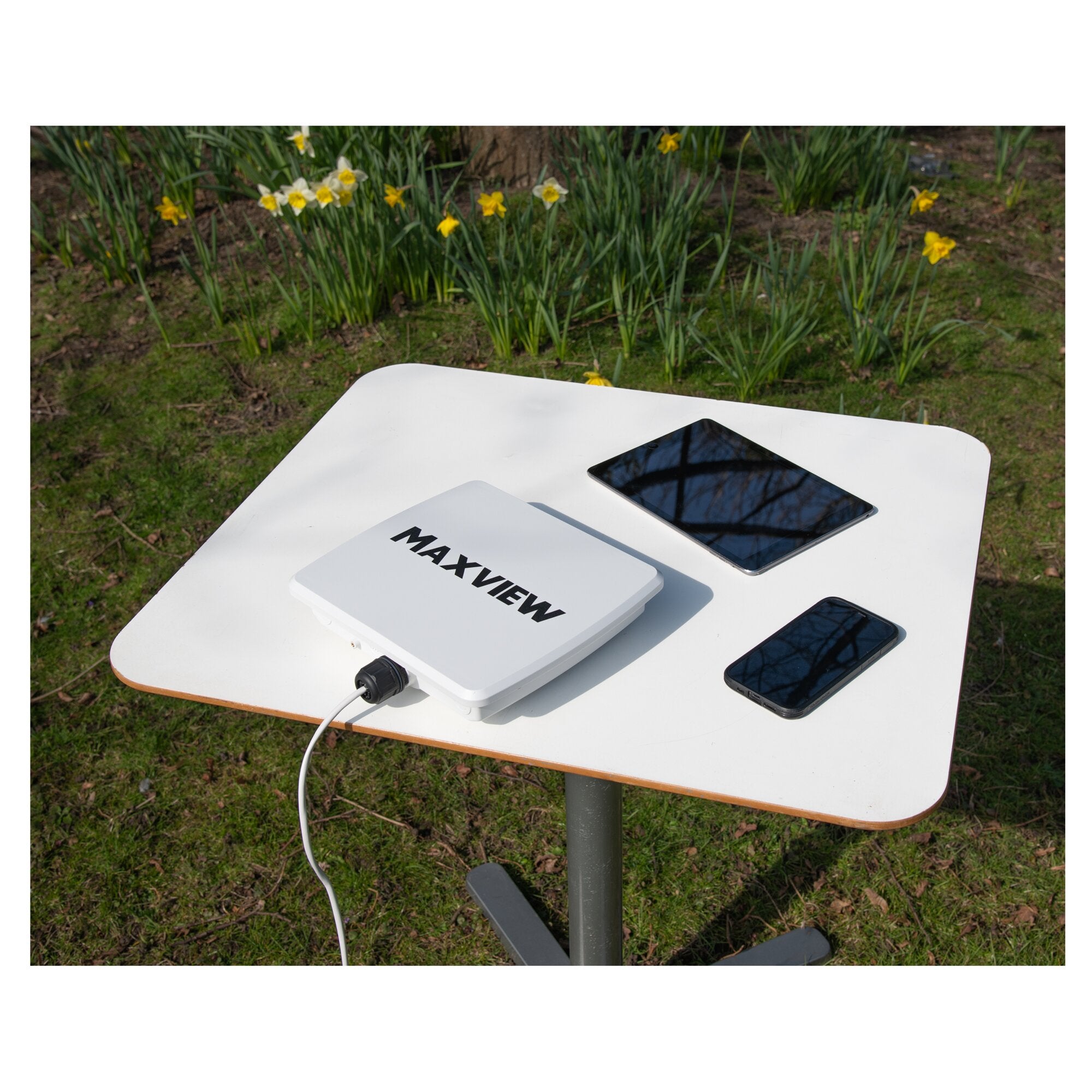 Außenbereich auf einem weißen Tisch mit einem MAXVIEW Roam Basecamp Router-Set, zwei Smartphones, Verpackung mit MAXVIEW-Logo, Kabel, Hintergrund Narzissen