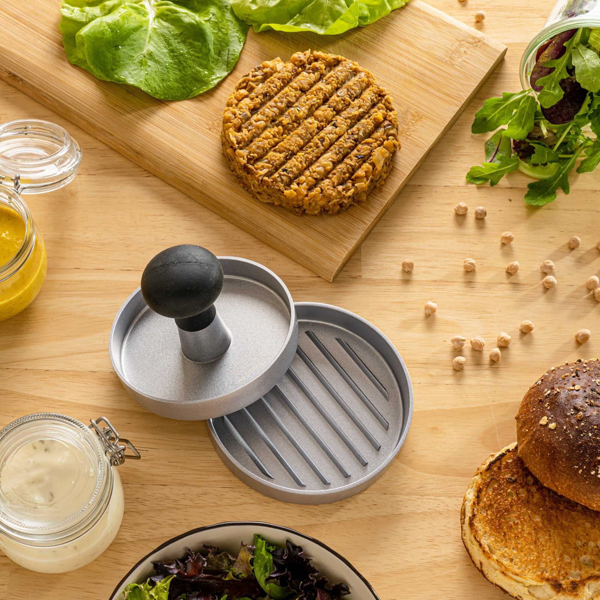 Hamburgerpresse aus Metall mit runder Form und Griff, daneben geformter Bratling auf Holzbrett, dazu frischer Salat, Brotlaib und Glas mit gelber Sauce
