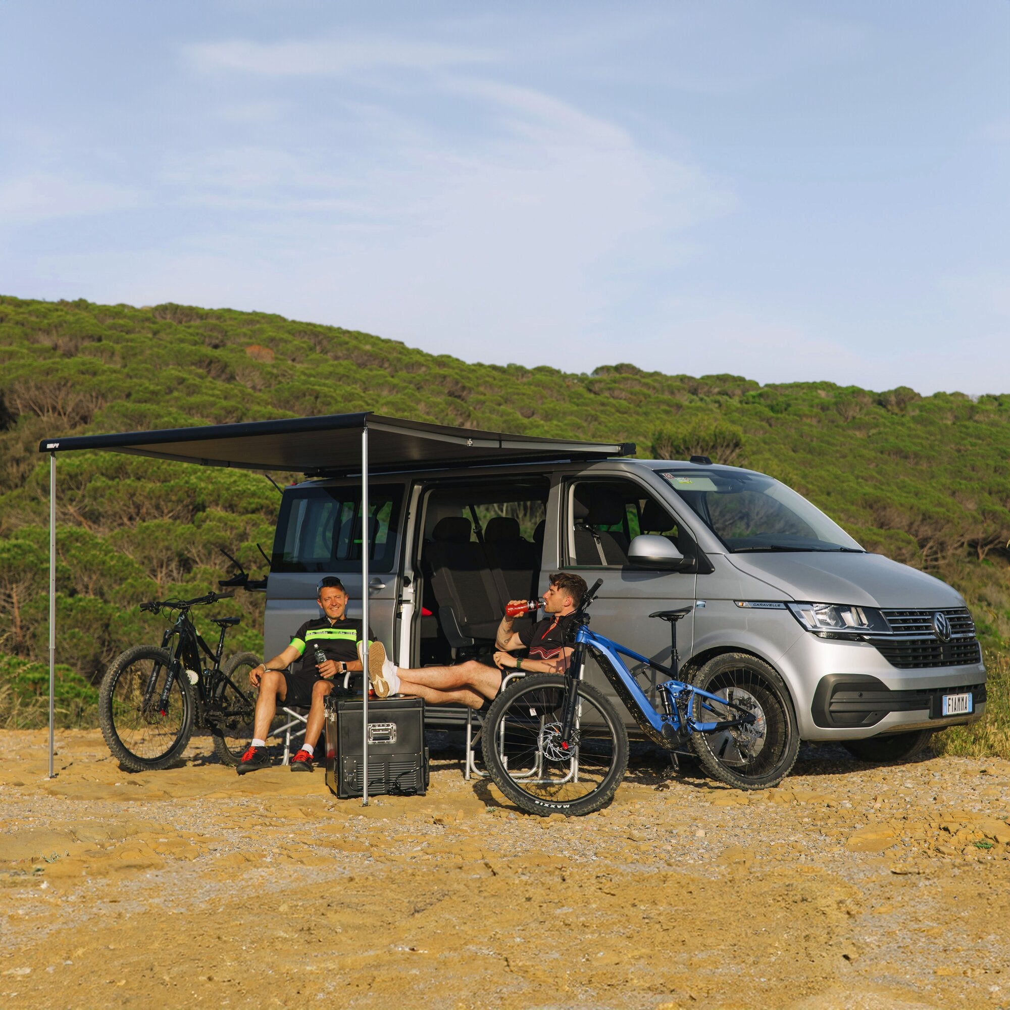 Markise F40van 270 cm am seitlichen grauen Camper ausgefahren; Stützen auf Kies, zwei Personen sitzen darunter, Fahrräder daneben Buschland