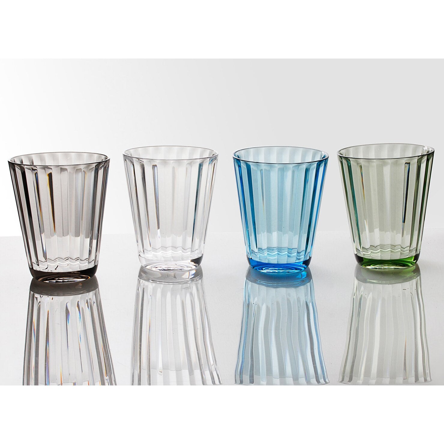 Vier farbige Trinkgläser mit vertikalen Rillen aus Glas in Braun, Klar, Blau und Grün stehen auf einem Tisch vor hellem Hintergrund