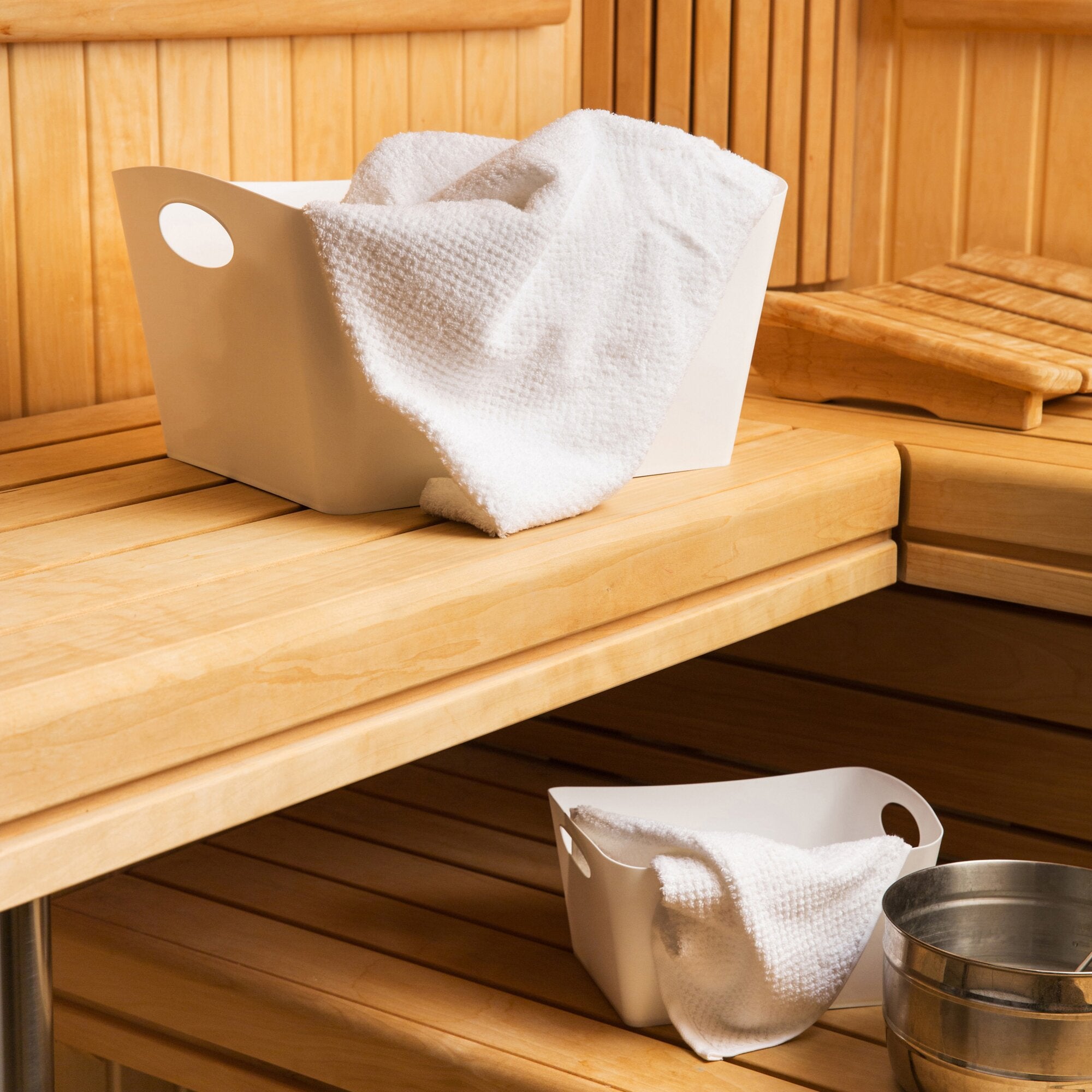 Sandfarbene Aufbewahrungsbox BOXXX M mit einem flauschigen weißen Handtuch drapiert, steht auf einer Holzbank in einer Sauna