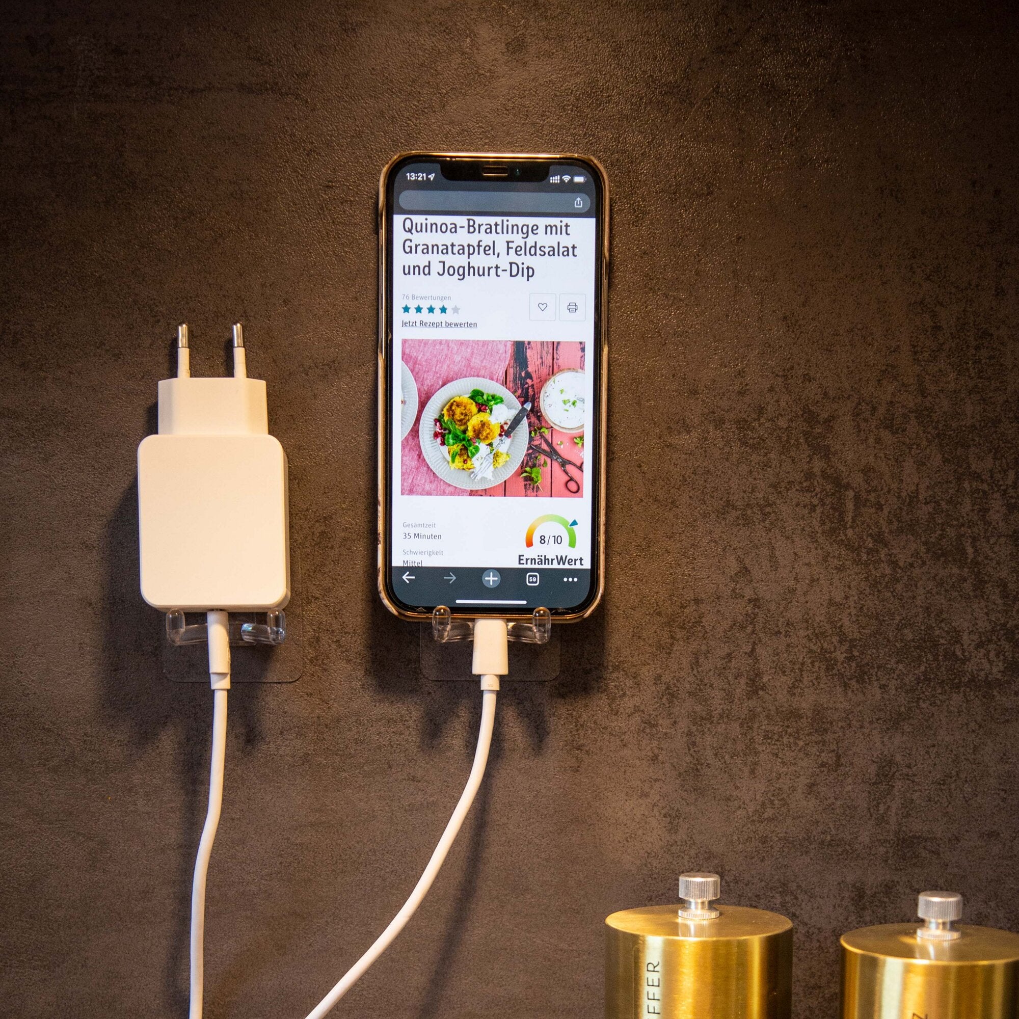 Smartphone mit Rezepttext Quinoa-Bratlinge mit Granatapfel, Feldsalat und Joghurt-Dip auf dunkler Oberfläche; links weißes Ladegerät, rechts zwei goldene Metallgewichte