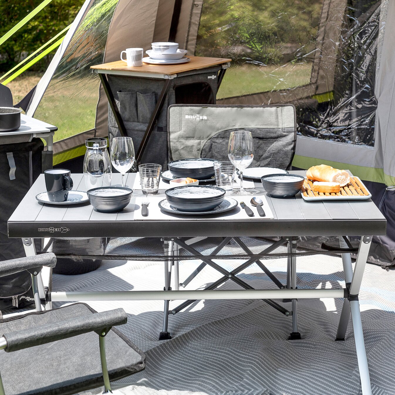 Rolltisch Accelerate im Campingsetup, graue Platte, Brunner-Logo, klappbare Beine, gedeckter Tisch mit Geschirr, Gläser, Besteck und Brot