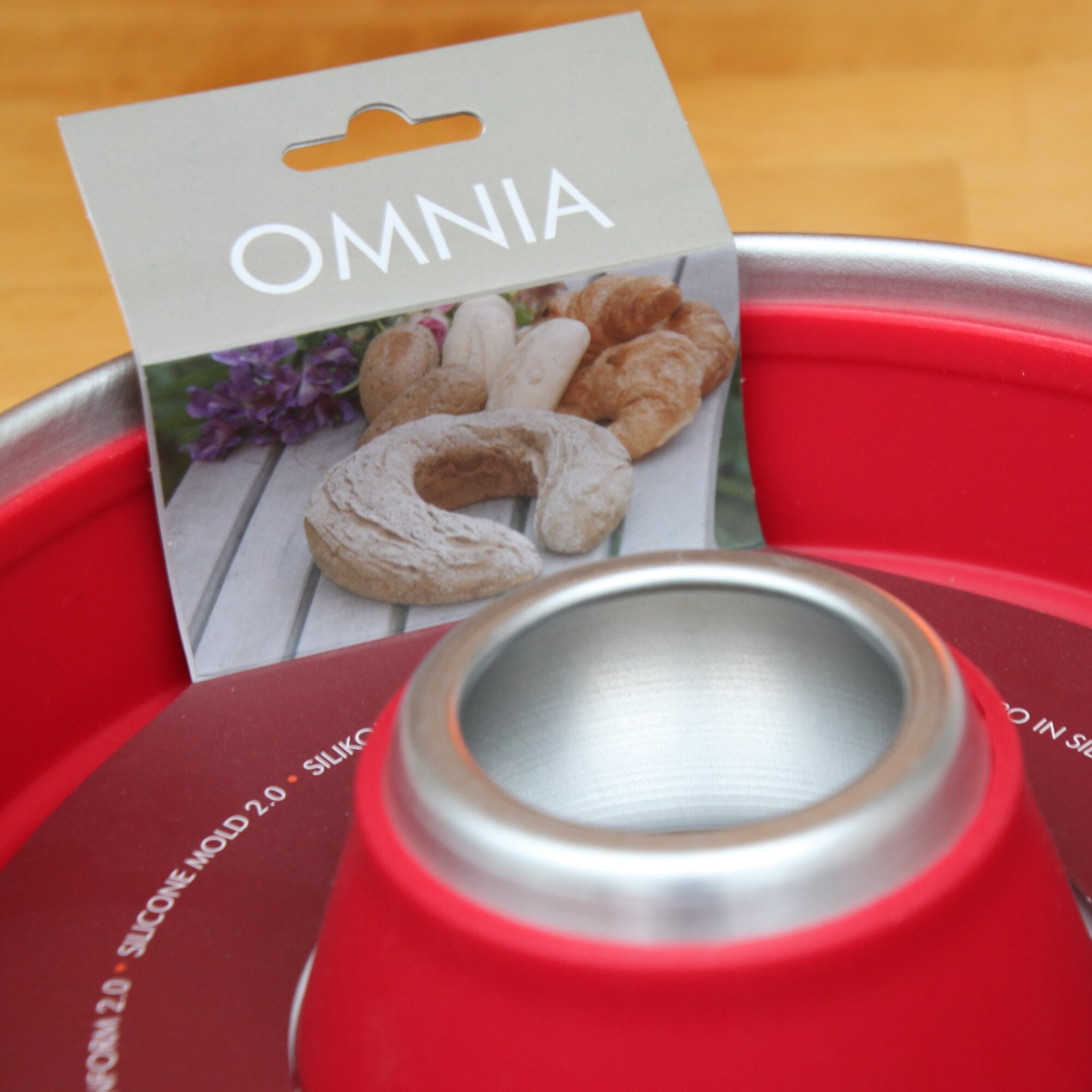 Muffin-Tab Backform aus Metall mit rotem Gehäuse und silberner Innenfläche, daneben eine Verpackung mit dem OMNIA-Logo