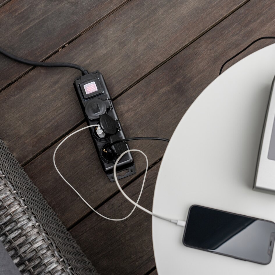 Schwarze 3-fach-Steckdosenleiste aus Kunststoff liegt auf Holzdielen; drei Steckdosen sichtbar, Kabel führt zu Smartphone am weißen Tisch
