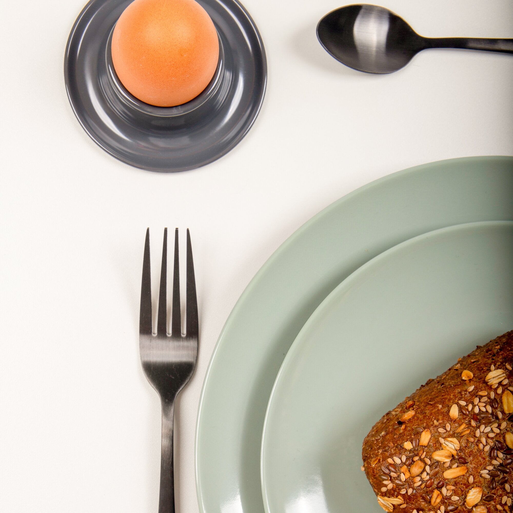 Gedeckter Tisch von oben mit hellgrünen Tellern, dunklem Teller darunter, orangefarbenem Ei, einer schwarzen Gabel und Brot mit Samen am Rand