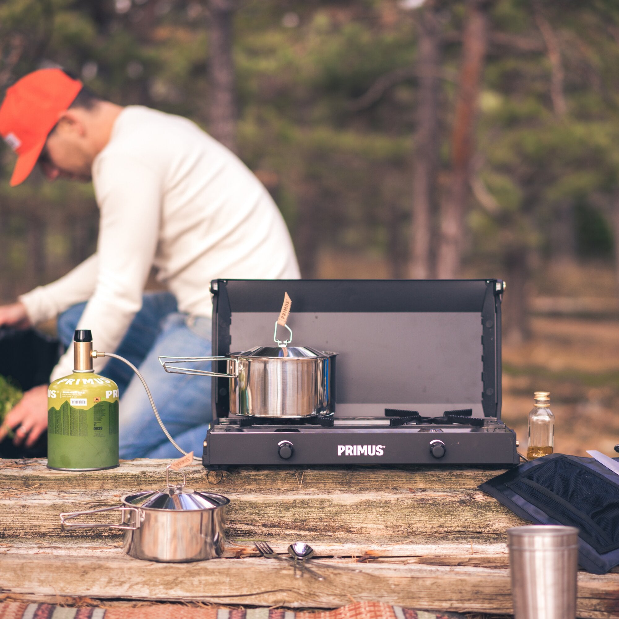 Portabler Gaskocher mit Edelstahltopf auf Holztisch, linke Propangasflasche, unscharfer Mann mit orangener Kappe im Hintergrund, Waldkulisse