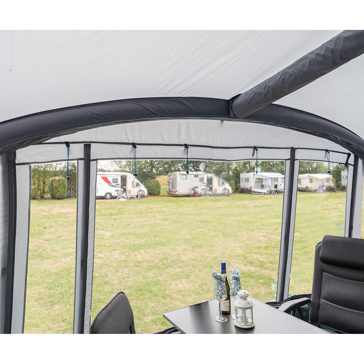 Innenansicht eines Dorado XL Teilzelts mit hellgrauer Dachplane, seitlichen Fenstern, gebogener Stange, Esstisch, Stühlen, Flaschen und Laterne; Campingplatz im Hintergrund